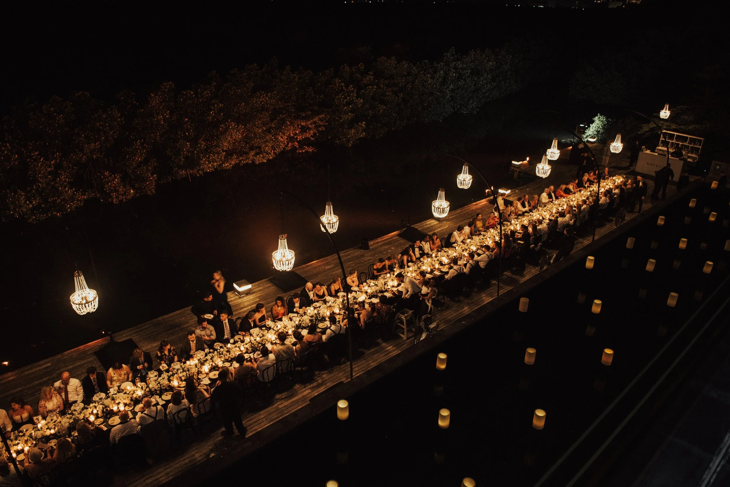 Wide shot overhead of a long table with wedding guest sitting at table. Chandeliers hang over the table