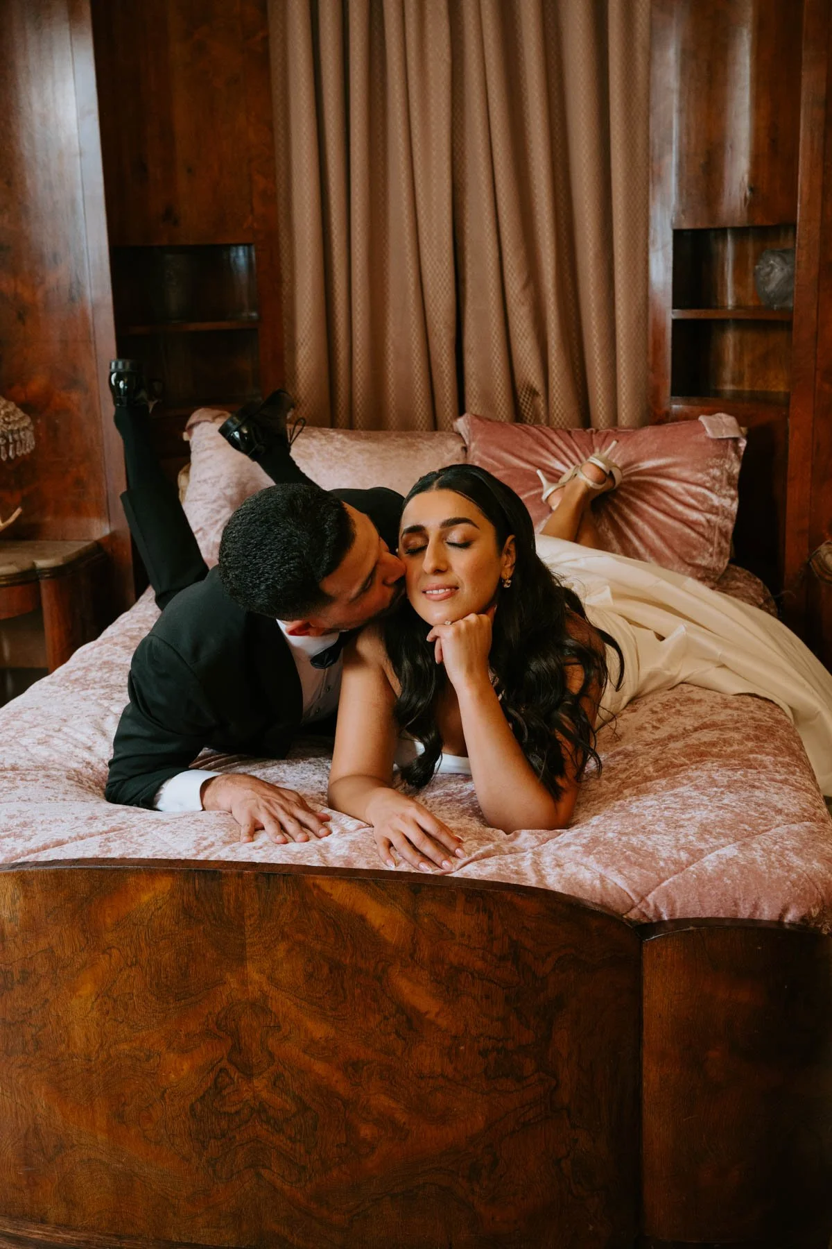 Bride and groom laying on their stomachs on a pink bed. Groom is leaning over kissing brides cheek.