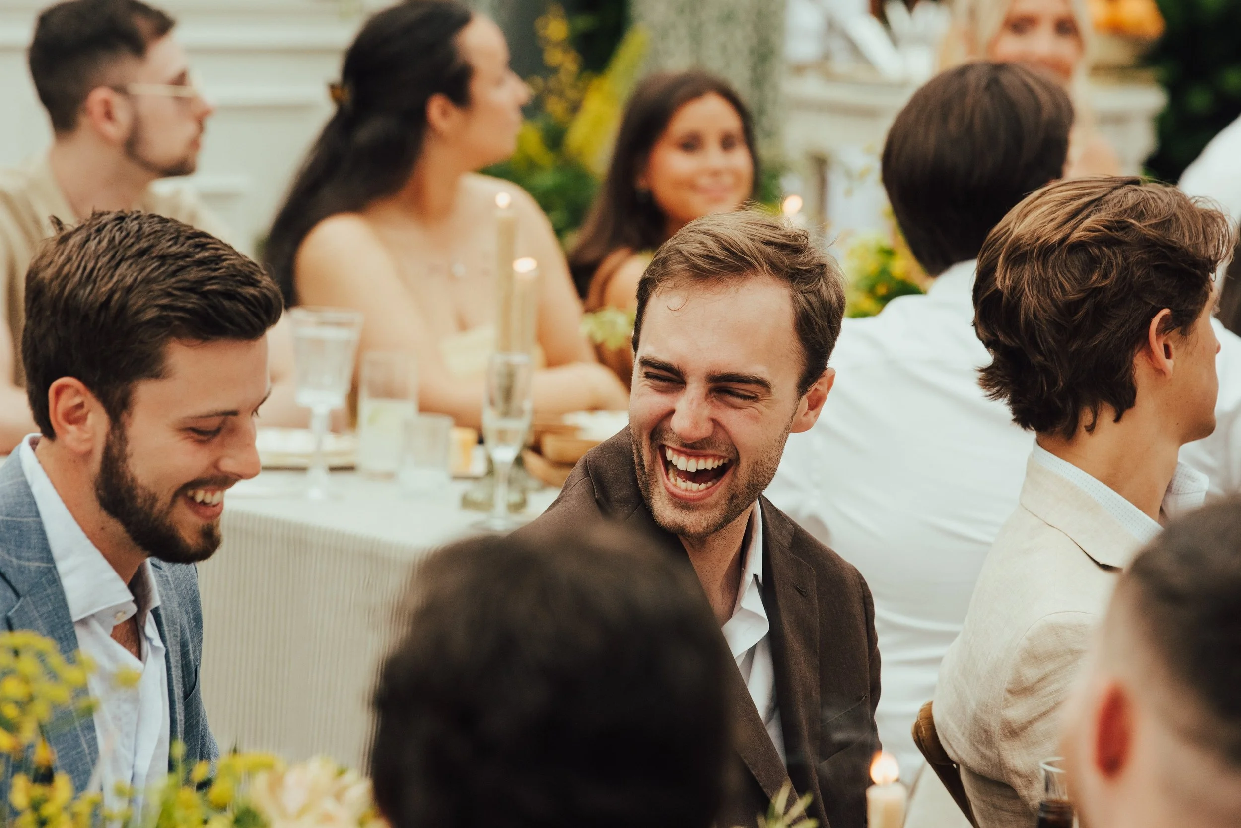 man in brown suit laughs sitting at a table of a wedding rehearsal dinner