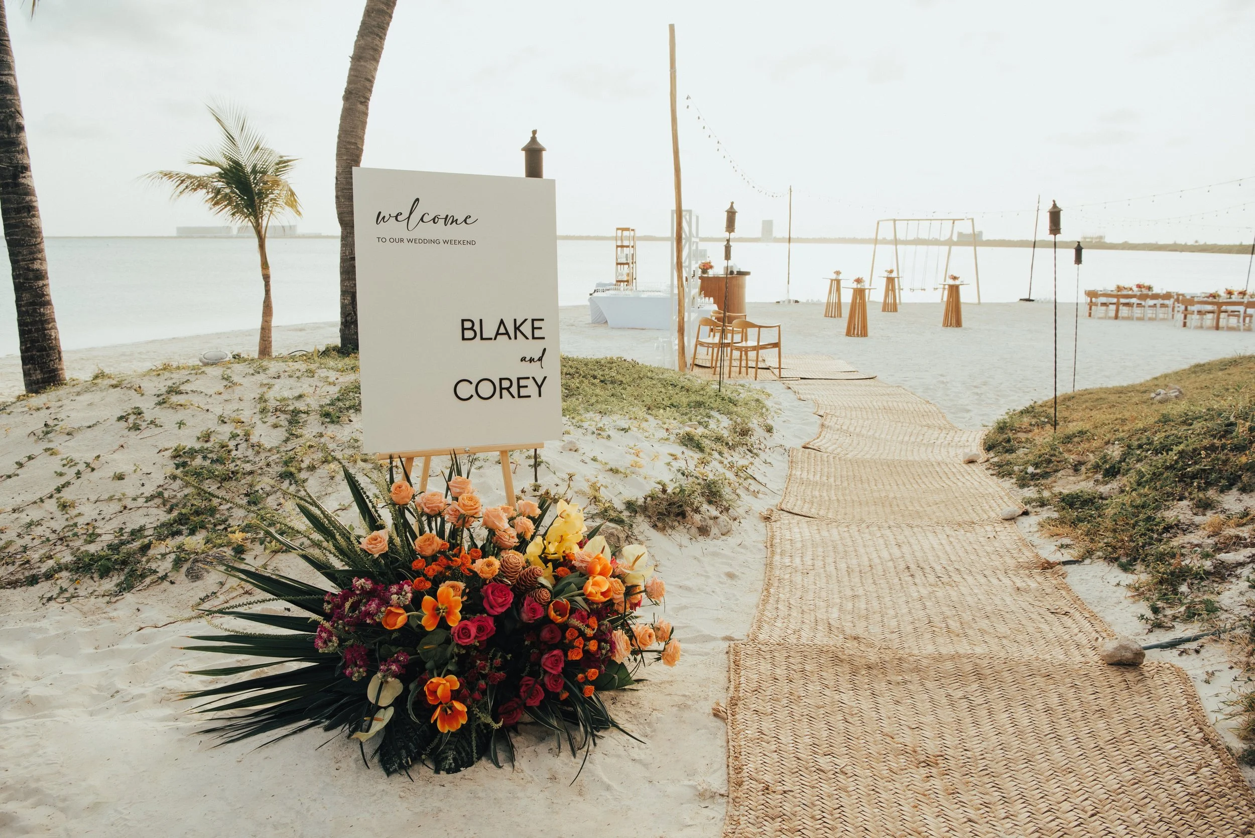 A wedding welcome sign with a floral arrangement in front of wedding welcome party set up on the beach