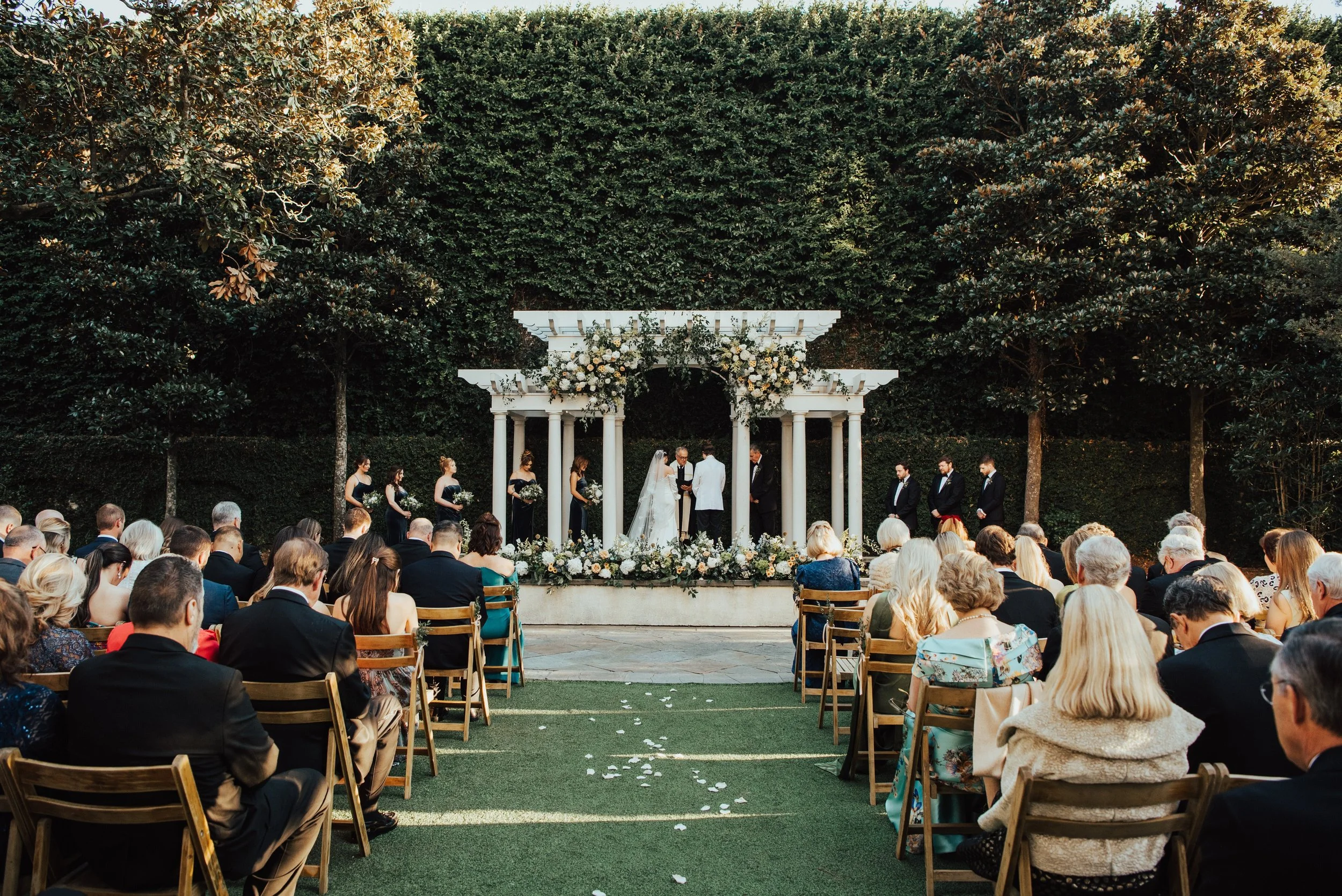 A wide shot of a wedding ceremony in progress. Ceremony is set against a wall of greenery with trees. There is a large white Arbor covered in flowers. You can see the backs of guests and the couple with their wedding party standing at the arbor.