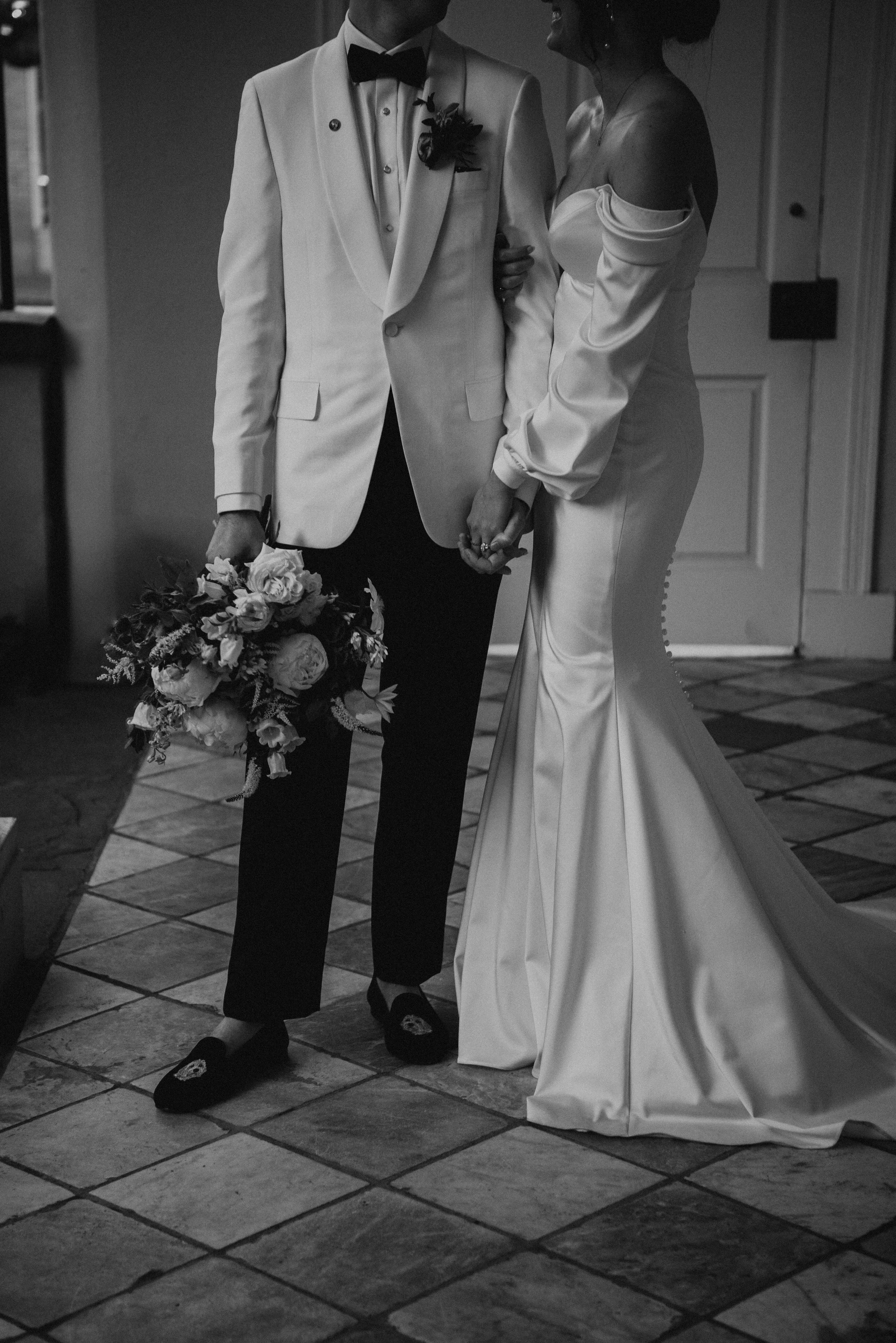 White and black photo of bride and groom from the neck down. Groom body is face camera as he holds the Brides bouquet by his leg. Bride's body is turned sideways to the camera as she hugs grooms arm