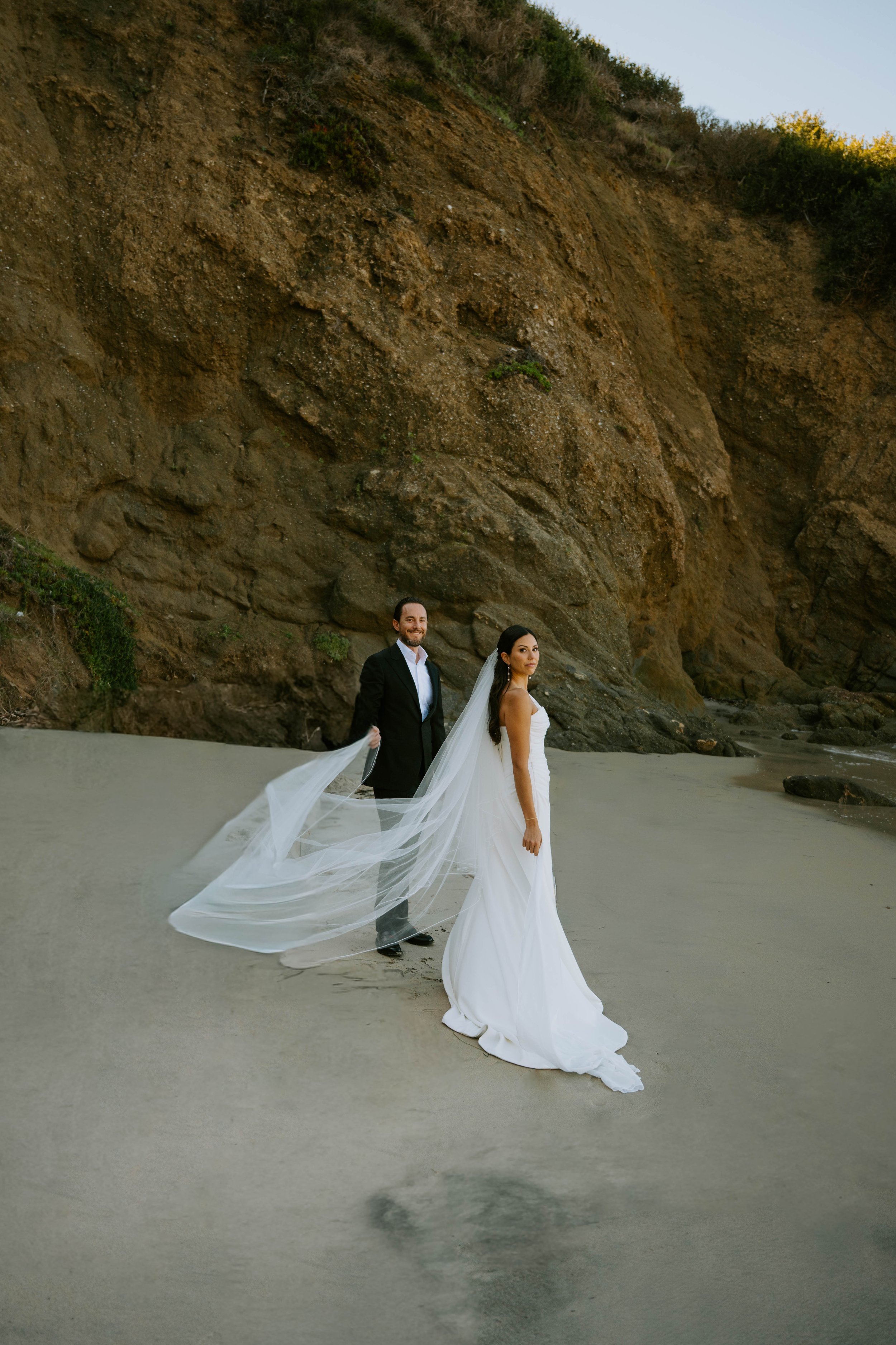 Bride and groom standing on beach in front of a cliff. both looking at the camera.