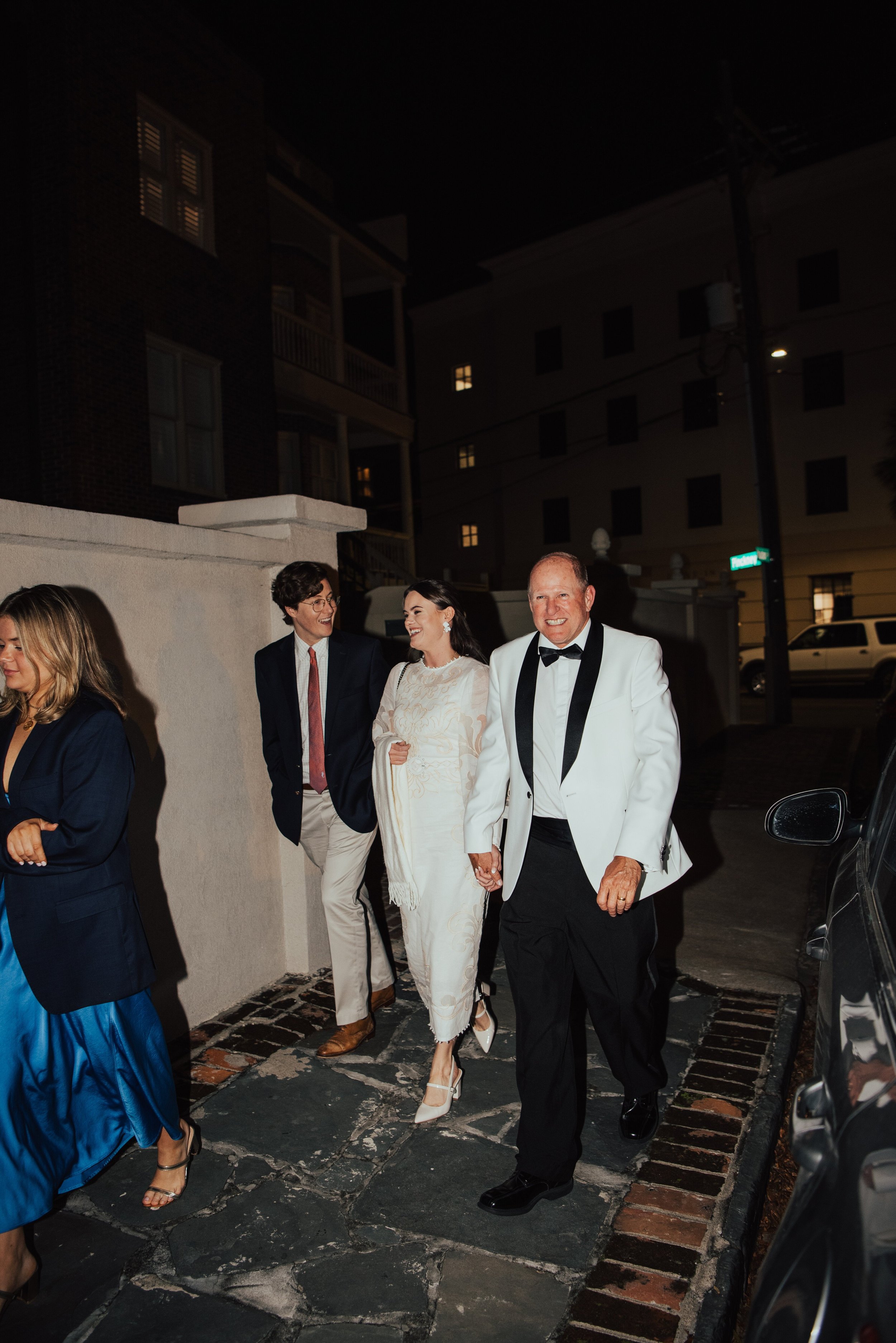 Bride walking down the street at night  holding her dad had and laughing with her fiancé