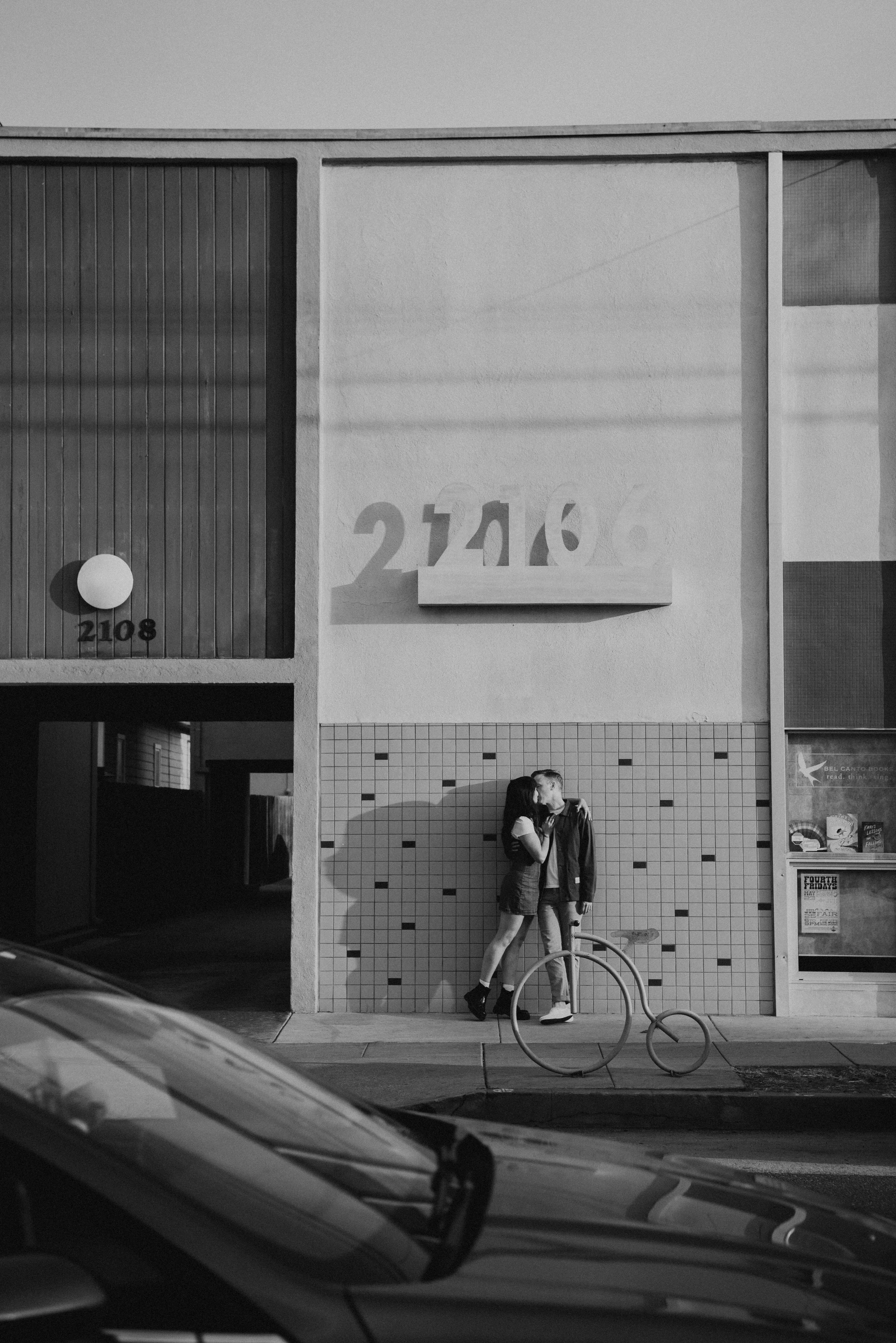 Black and white photo of wide shot of couple kissing while standing on the side walk as car passes in the foreground