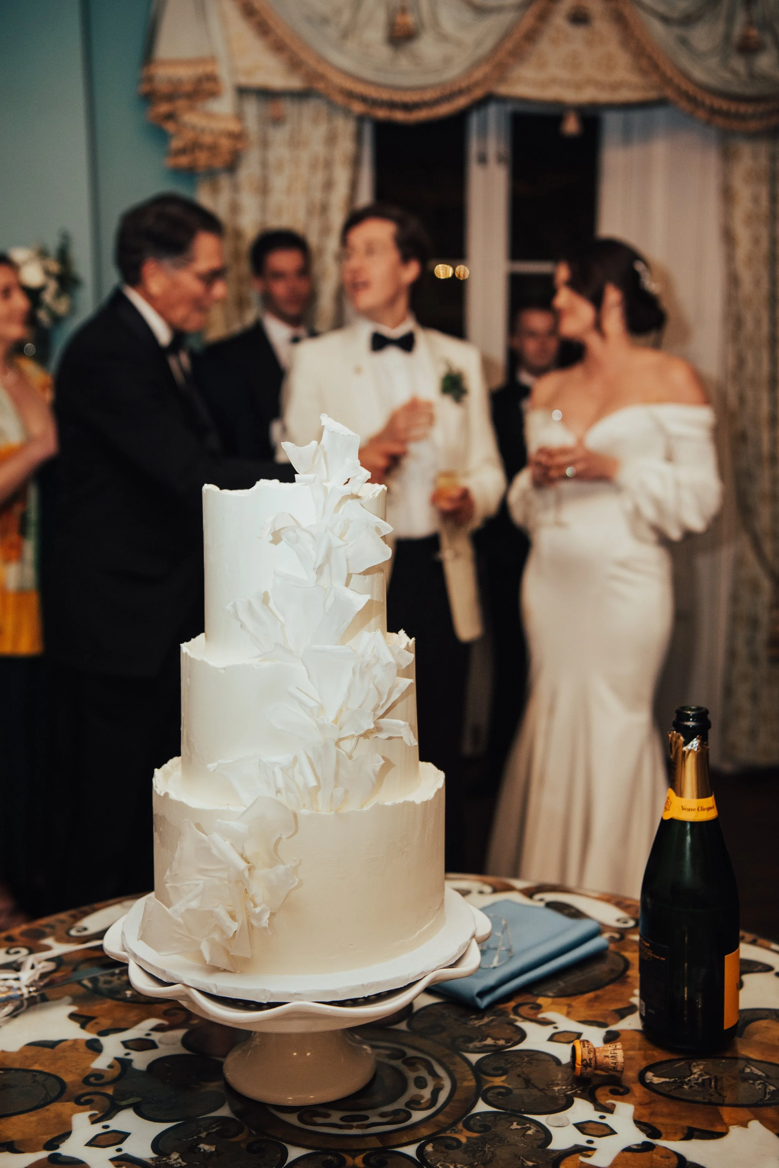 All white wedding cake in focus next to an open bottle of champagne. You can see the bride and groom in the background talking with guest