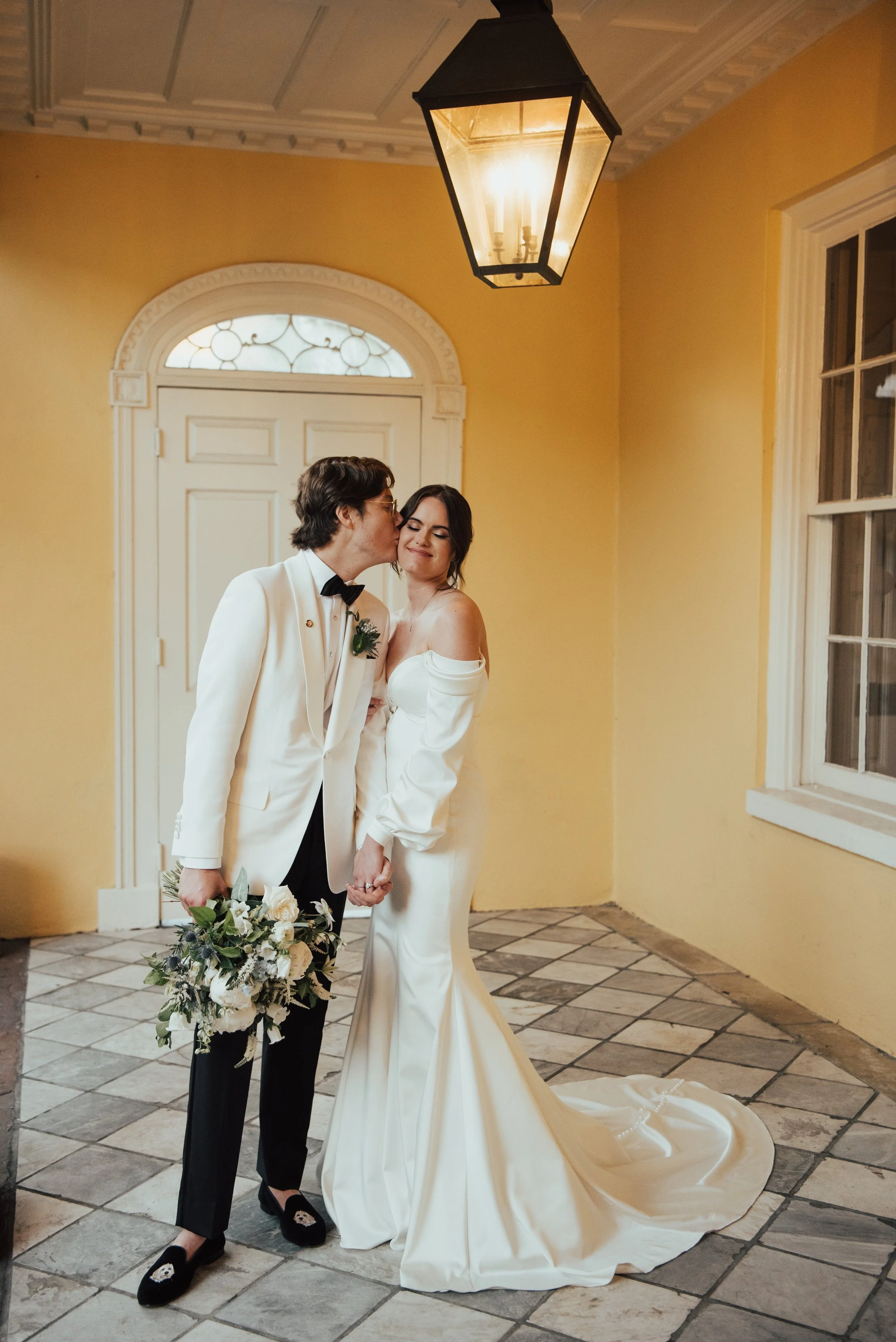 Bride and groom standing in front of yellow walls. Groom is kissing brides cheek and holding her bouquet. Bride is smiling with her eye closed and hugging the grooms arm