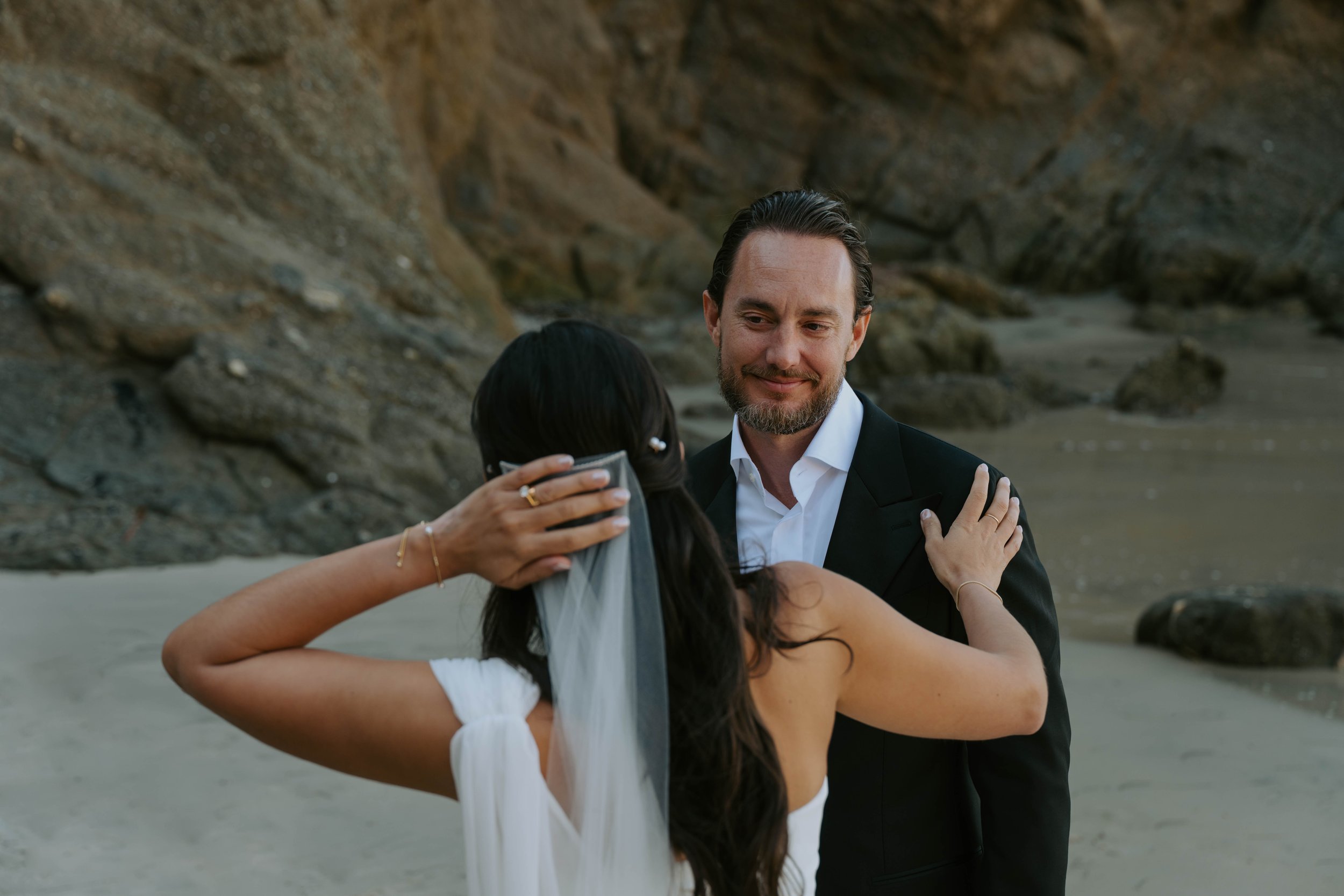 the back of the bride to the camera holding her veil on her head. Groom is facing camera looking at camera smiling.