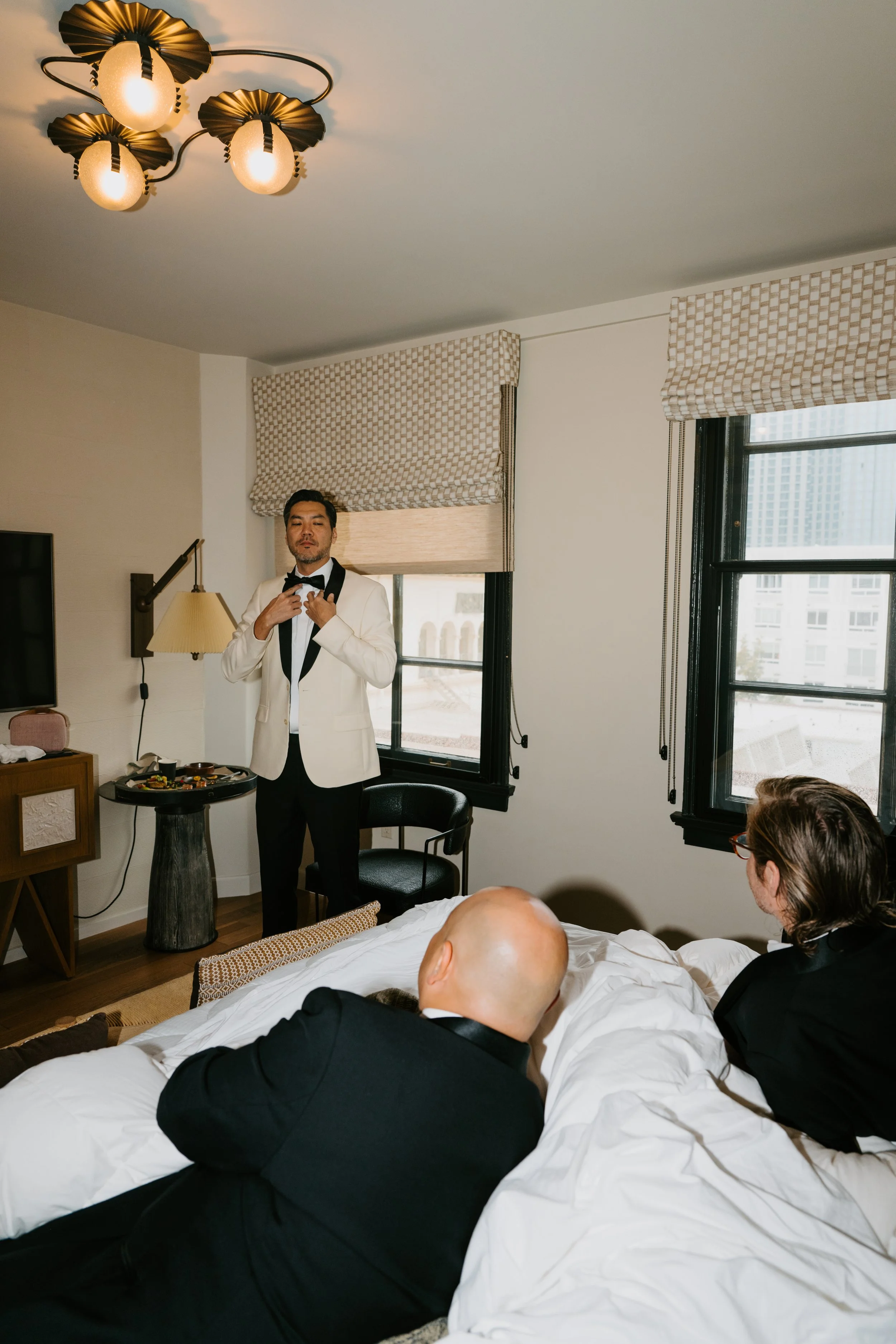 Groom fixing his tie and two groomsmen lay on the bed in tuxedos watching