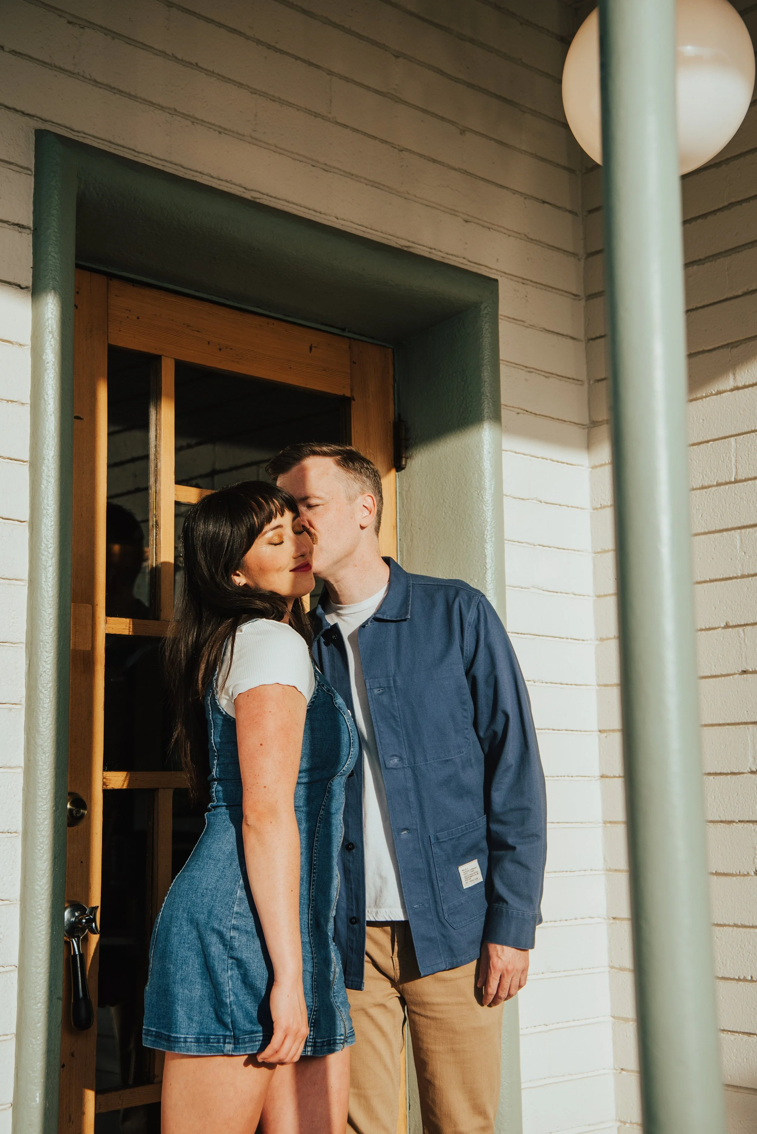 couple standing in front of a door as the sun shines on them. Guys is kissing girls cheek