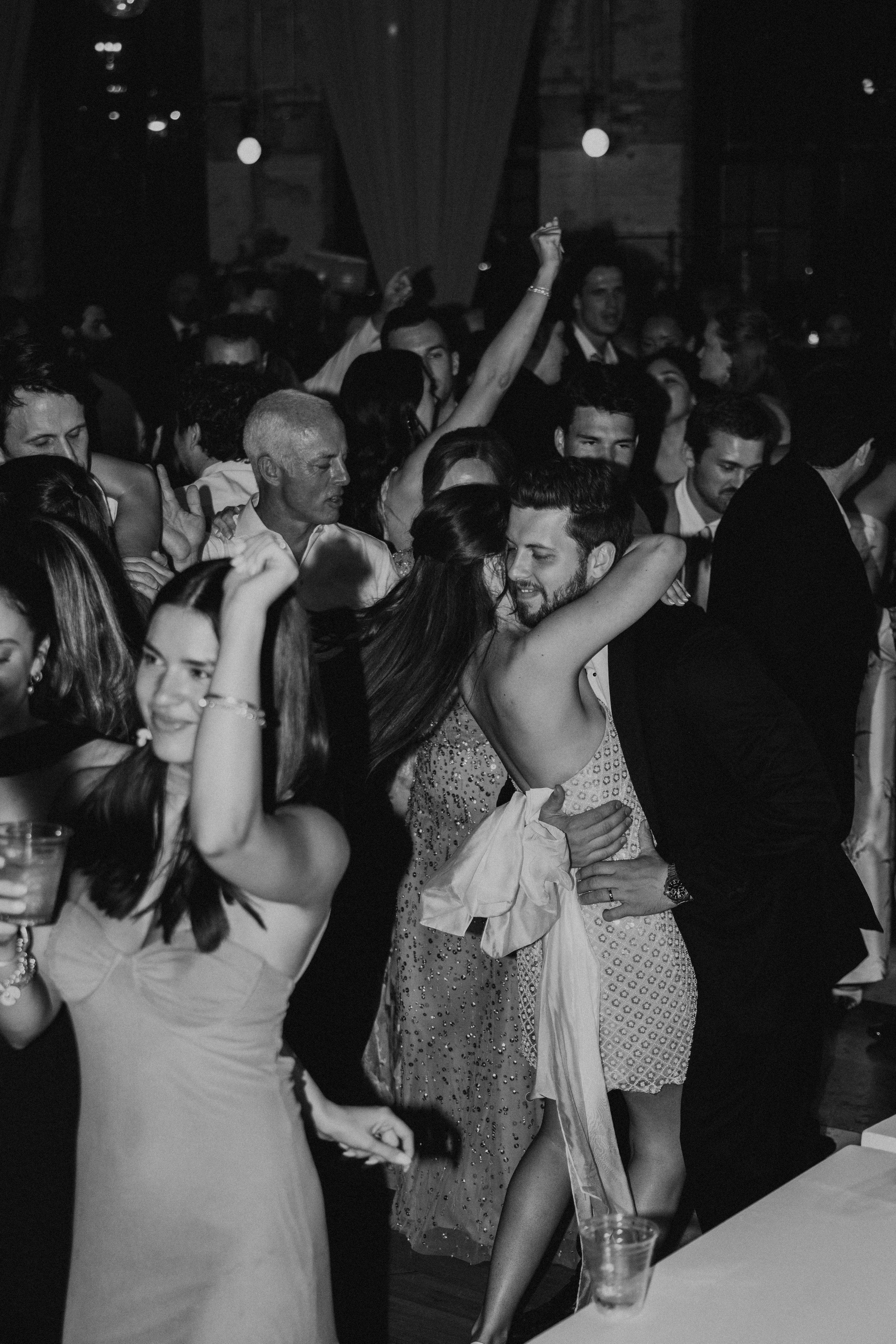 Black and white photo of bride and groom tightly hugging as the dance on the dance floor with the guest all around