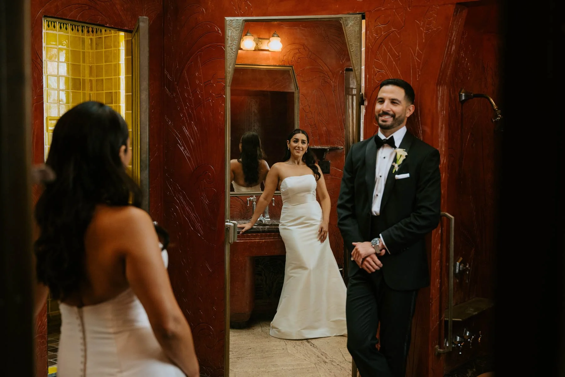 Bride and groom in red bathroom. They are standing opposite each other. Bride is seen in mirror door next to groom and groom is facing camera.