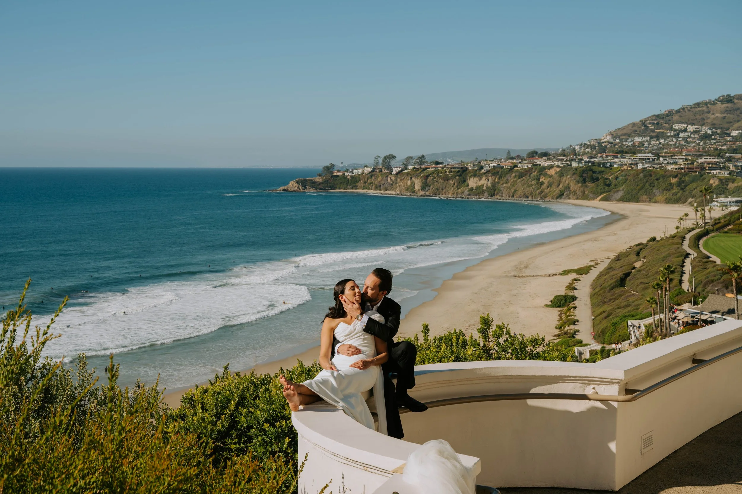 Bride and groom sitting on a wall with the ocean and land in the background