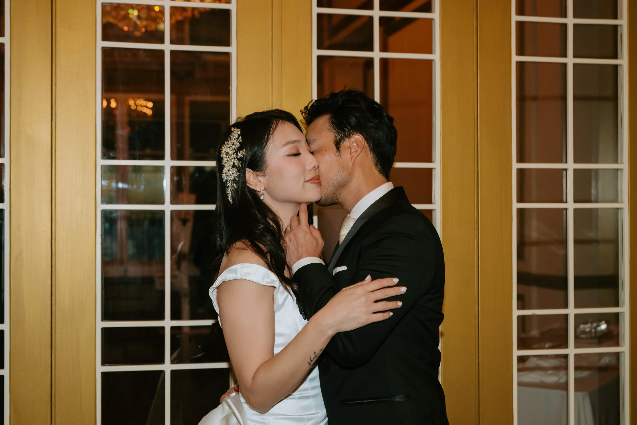 Bride and groom standing in front of gold frame doors with window that lead to a ballroom. Groom is whispering in Brides ear