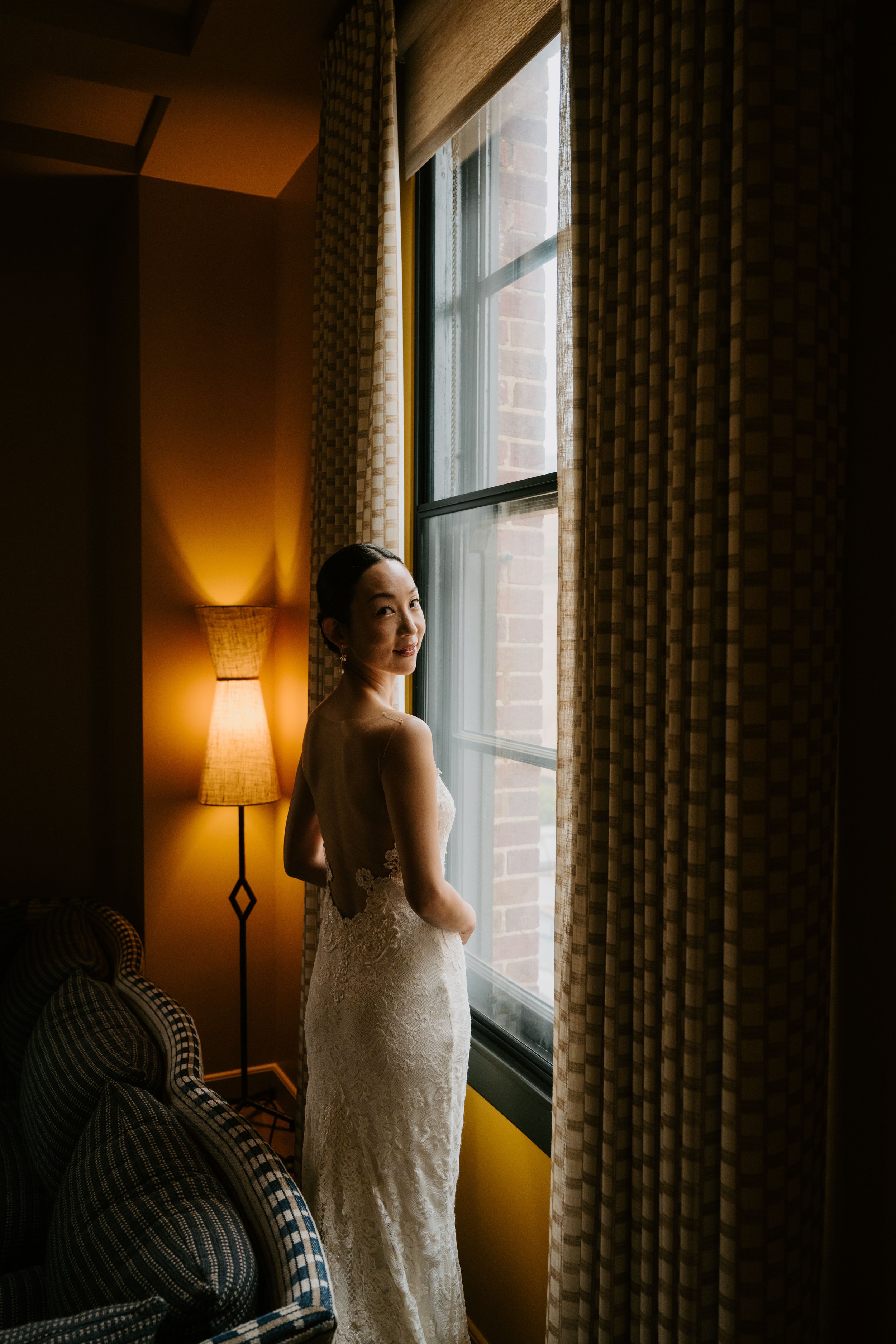 Bride standing at the window in a dark room looking back over her shoulder at the camera