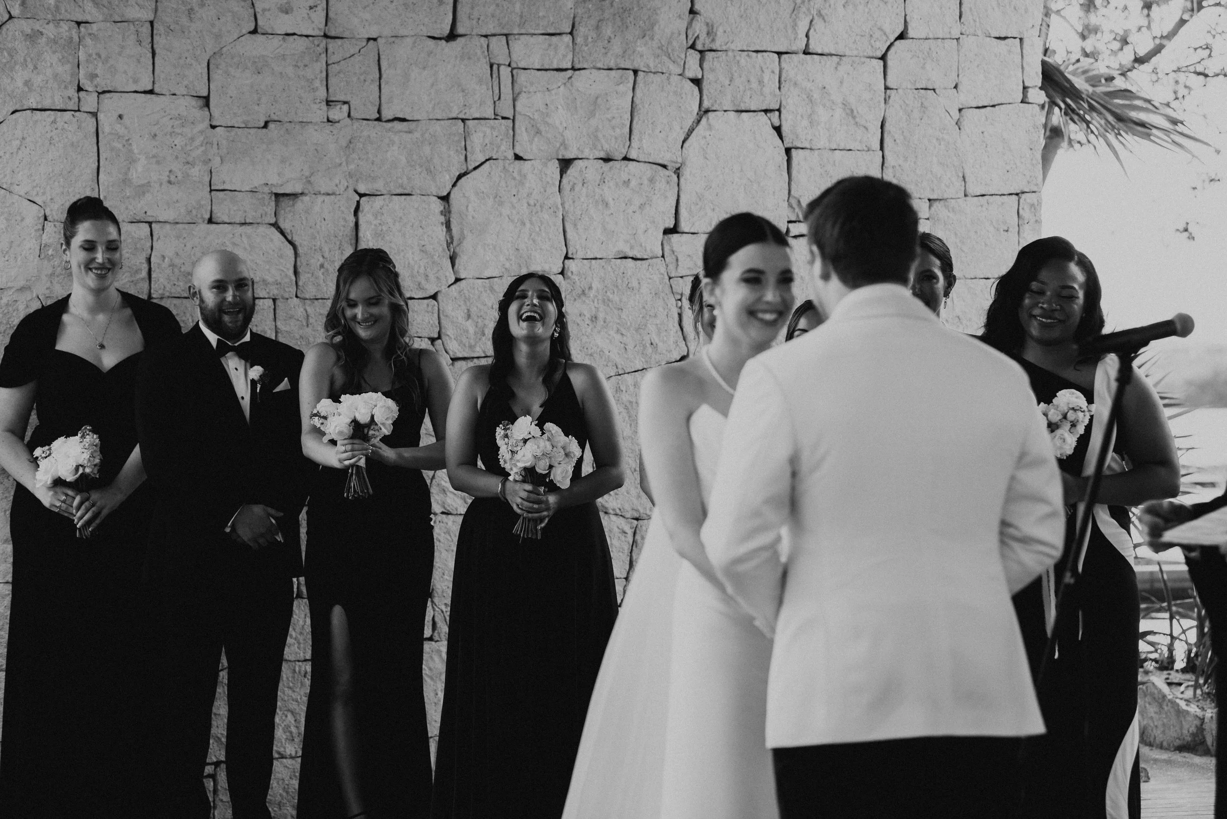 black and white photo of wedding party laughing behind couple during the wedding ceremony