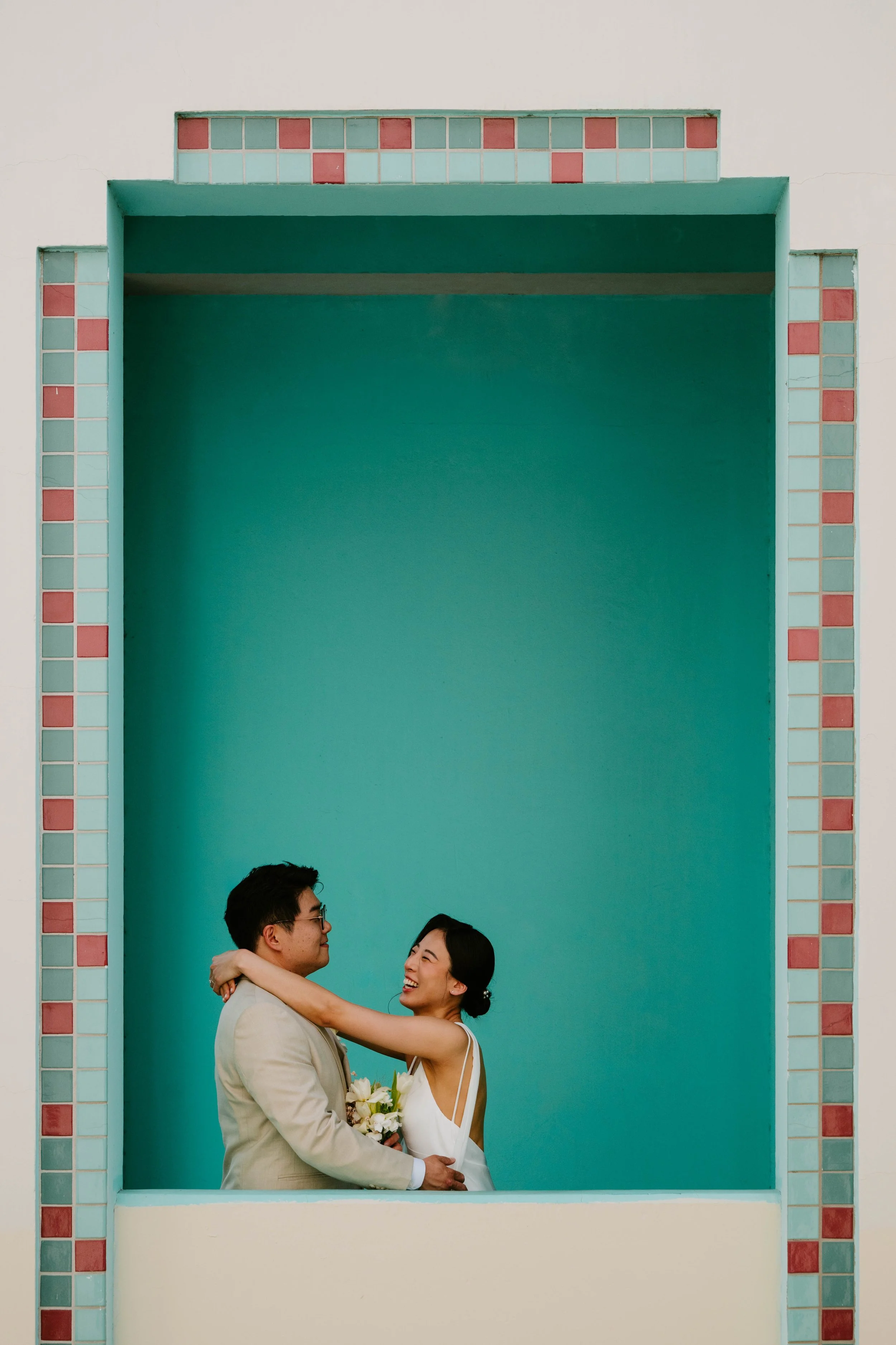 Bride and groom seen through a window. Bride is laughing with her arms around the grooms neck