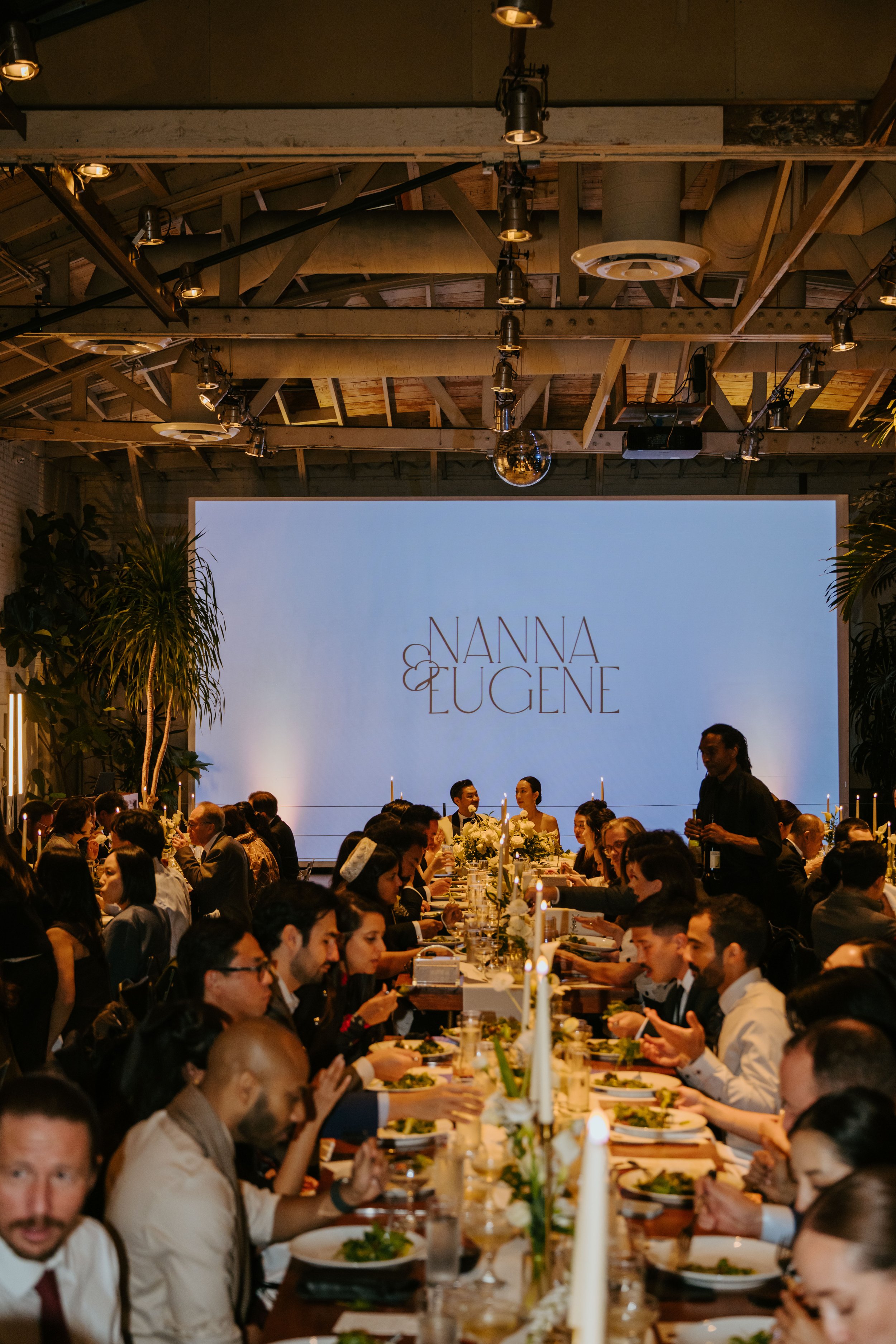 Photo of wedding guest eating at a long table. Focus is on the large screen behind them that has the bride and groom's name on it. Nanna & Eugene