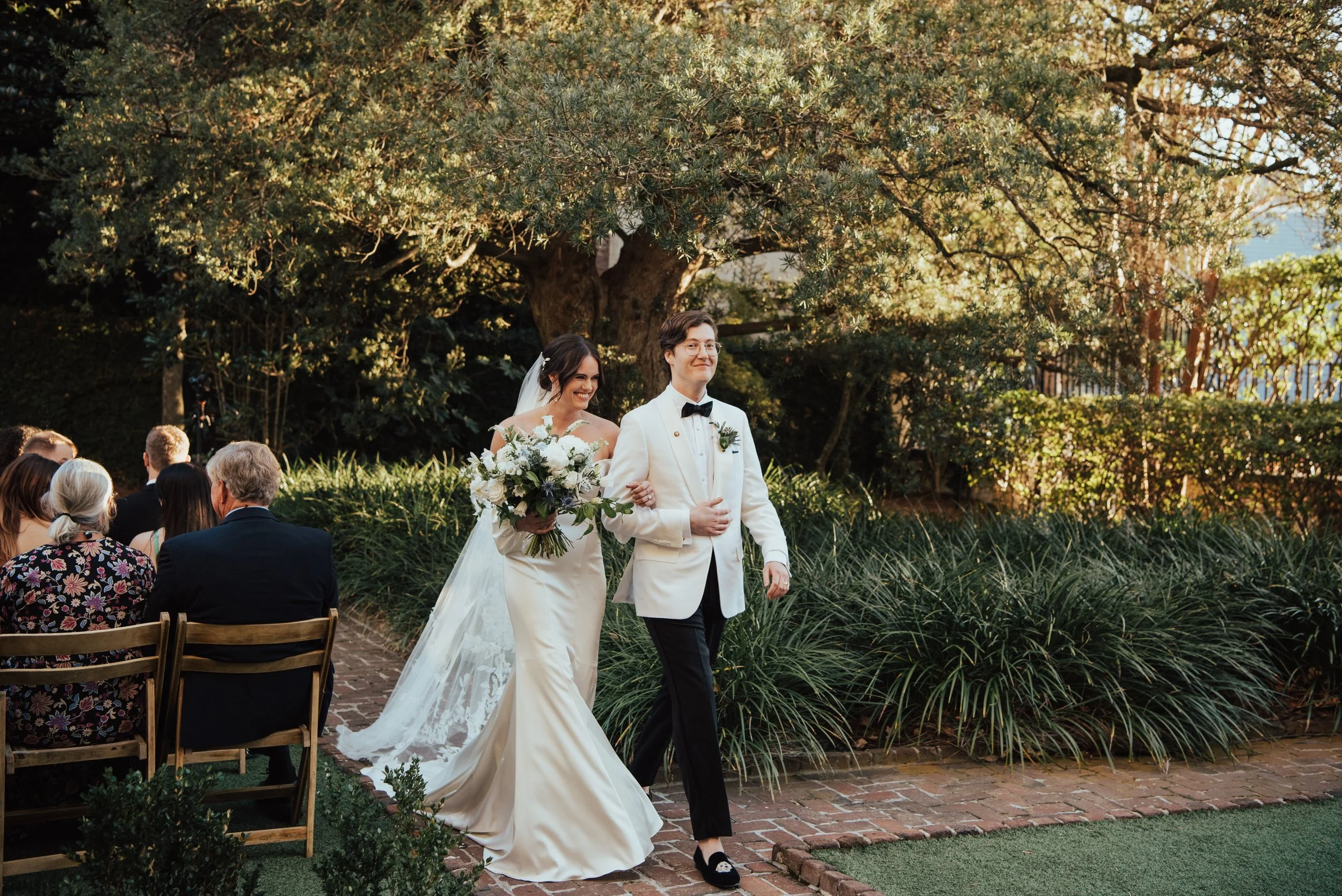 Bride and Groom in a white tuxedo jacket walking back down the aisle after their ceremony. Both are smile as Bride hugs grooms arm