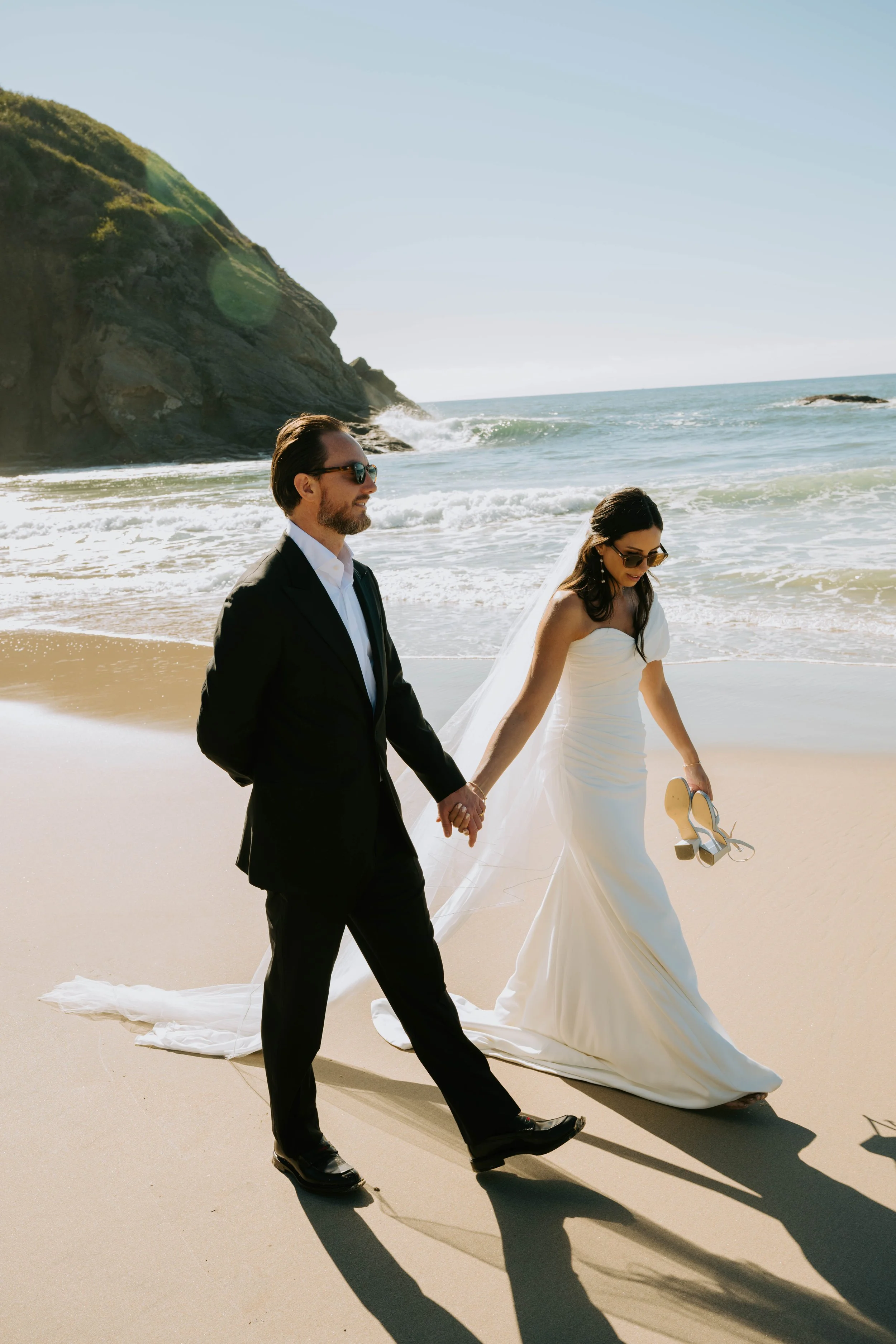 Bride and groom walking on the beach holding hands while wearing sunglasses