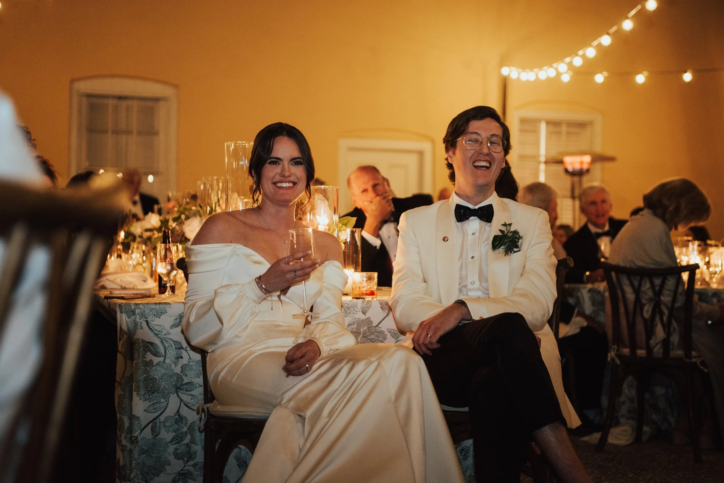 Photo of birde and groom sitting in front of their table smiling. The backdrop is the wall of a yellow house and their are bistro lights overhead.