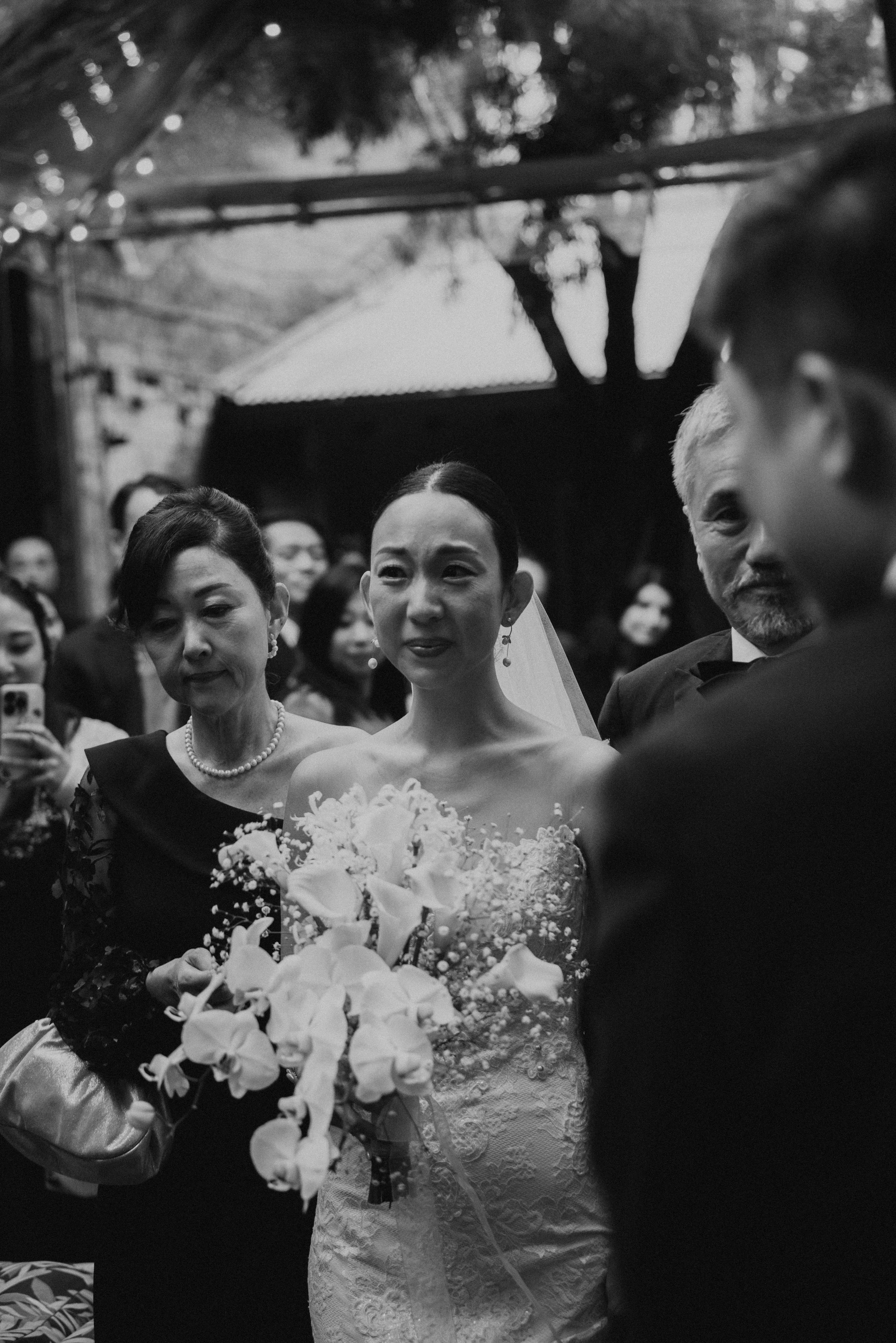 black and white photo of bride tearing up as she walks down with aisle with her mom and dad