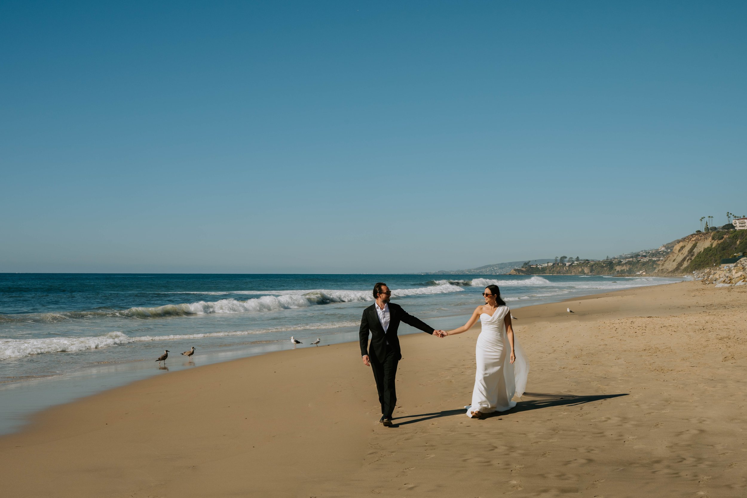 Bride and groom walking on the beach holding hands with blue sky, ocean and coastline in the background