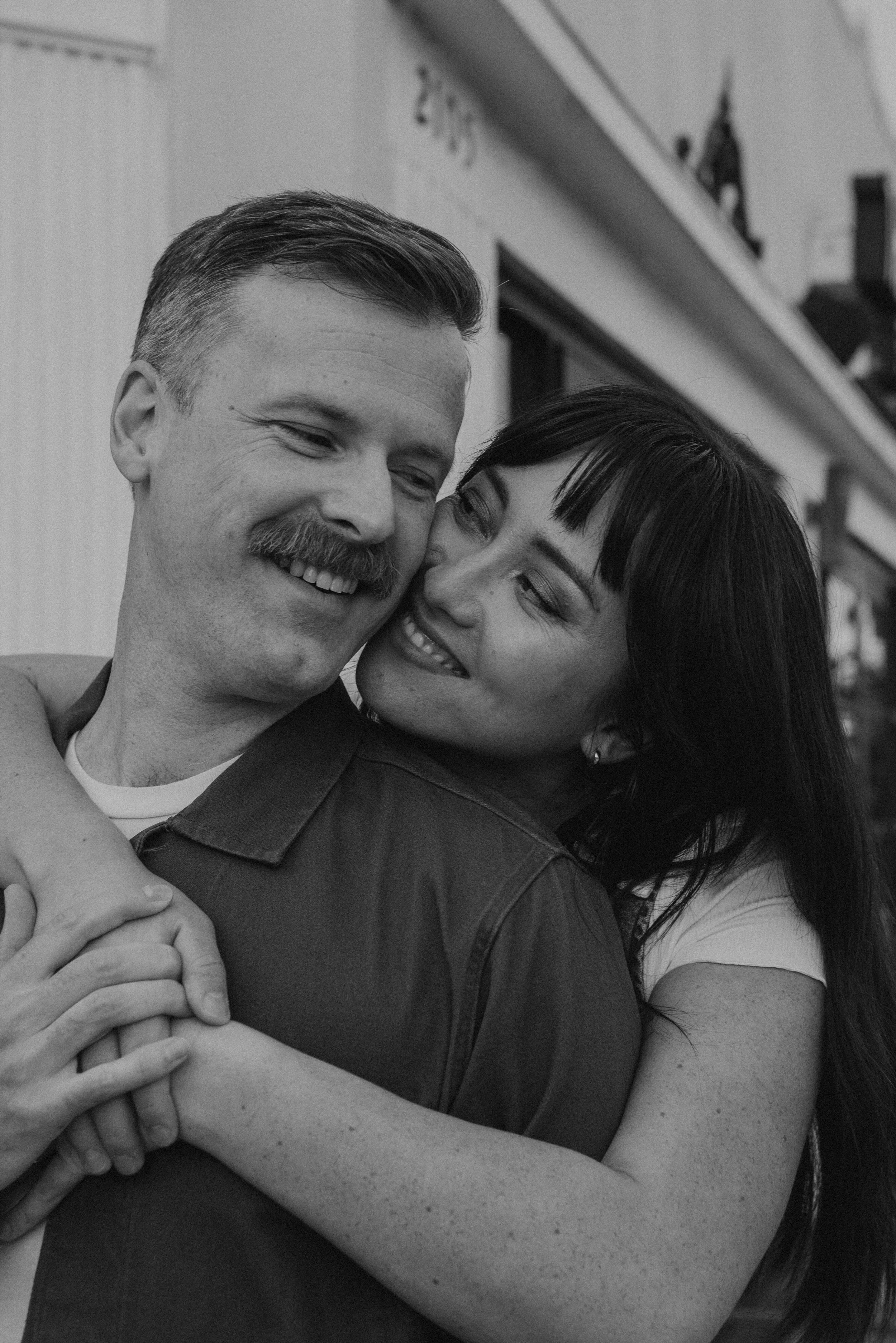 Black and white photo of girl hugging guy from behind. They are both smiling with their cheeks touching