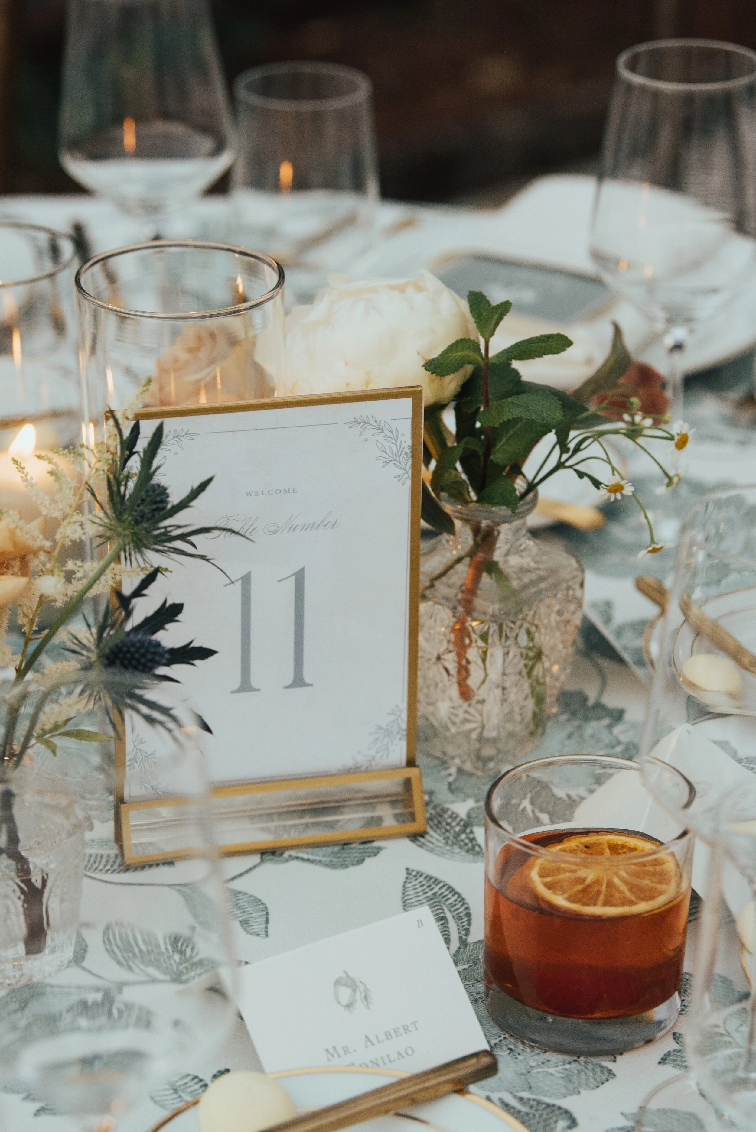 close up shot of wedding table number surrounded my florals. A cocktail is sitting on the table in front