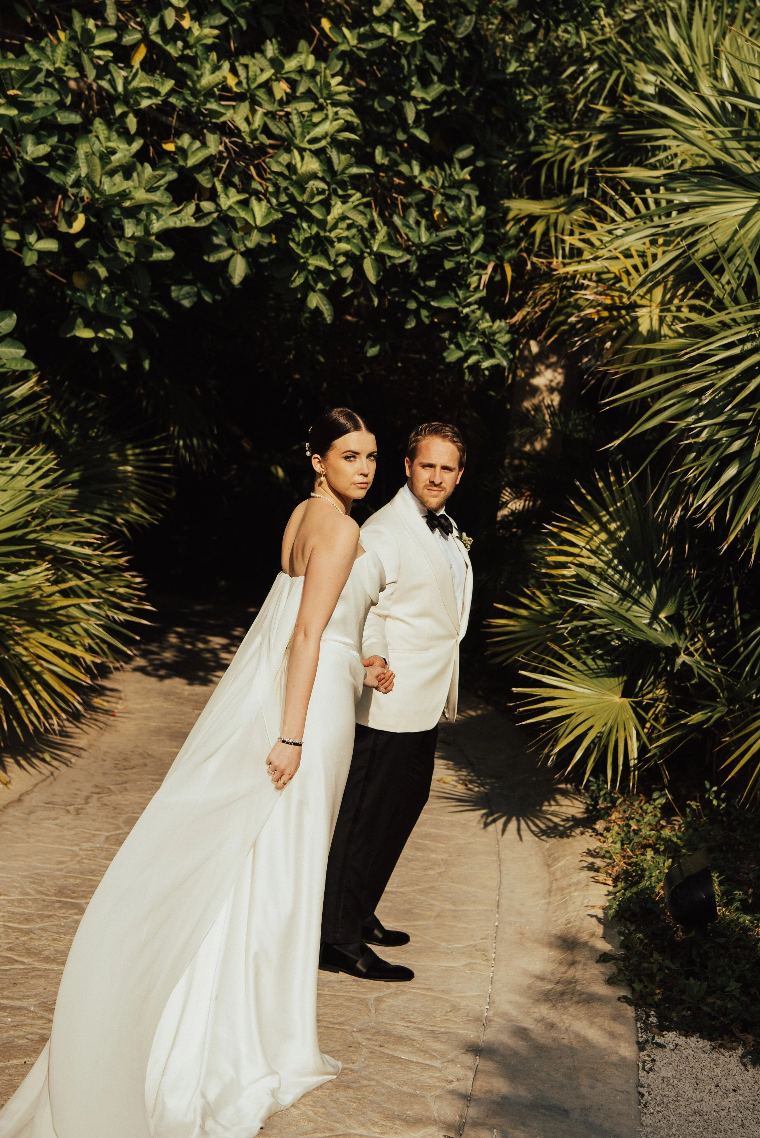 bride and groom walking away from the camera. Bride is looking back over her shoulder at camera. groom looking back at her. The couple is surrounded by greenery