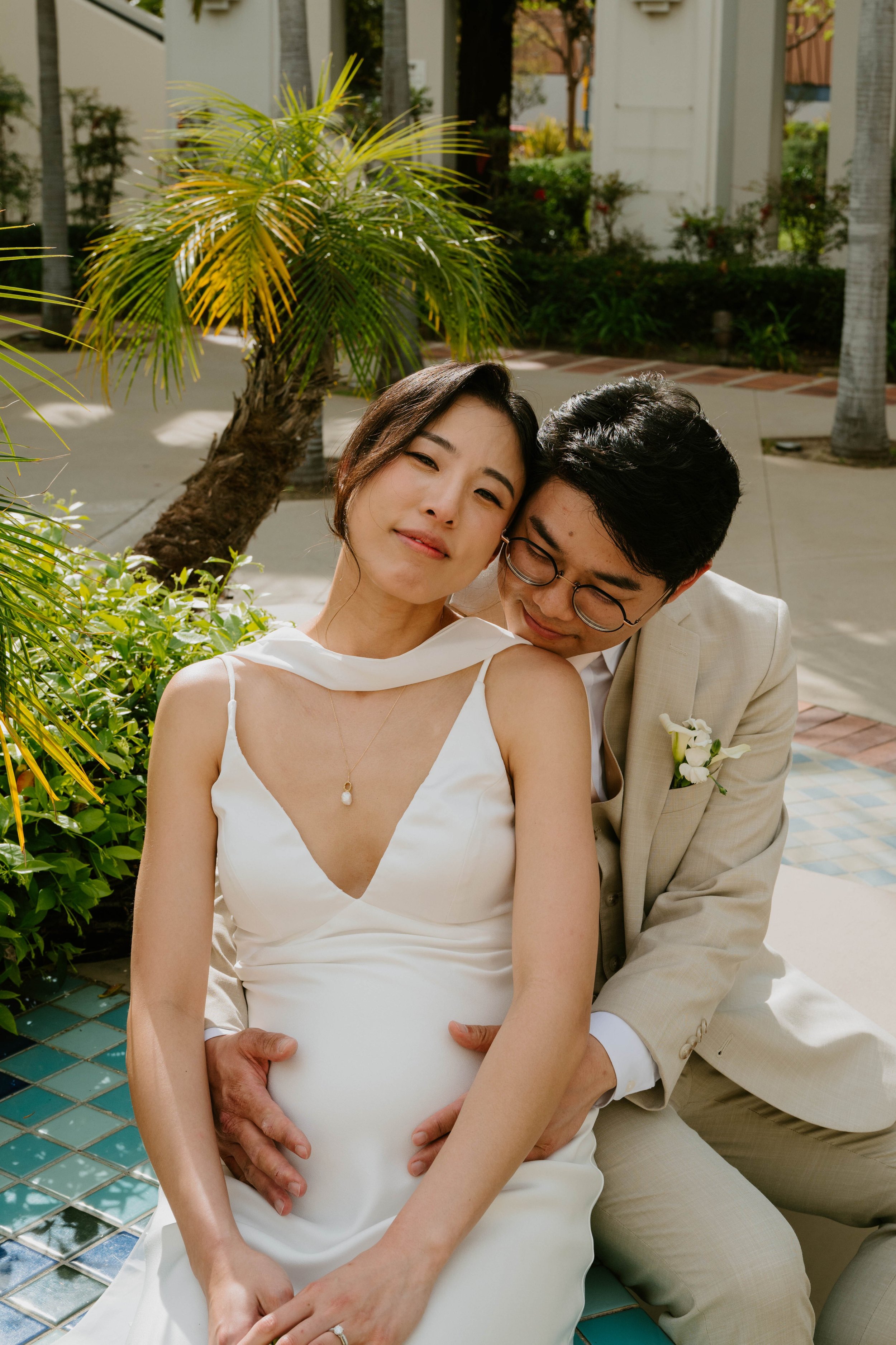 Groom hugging bride from behind and holding her baby bump as bride looks at the camera.
