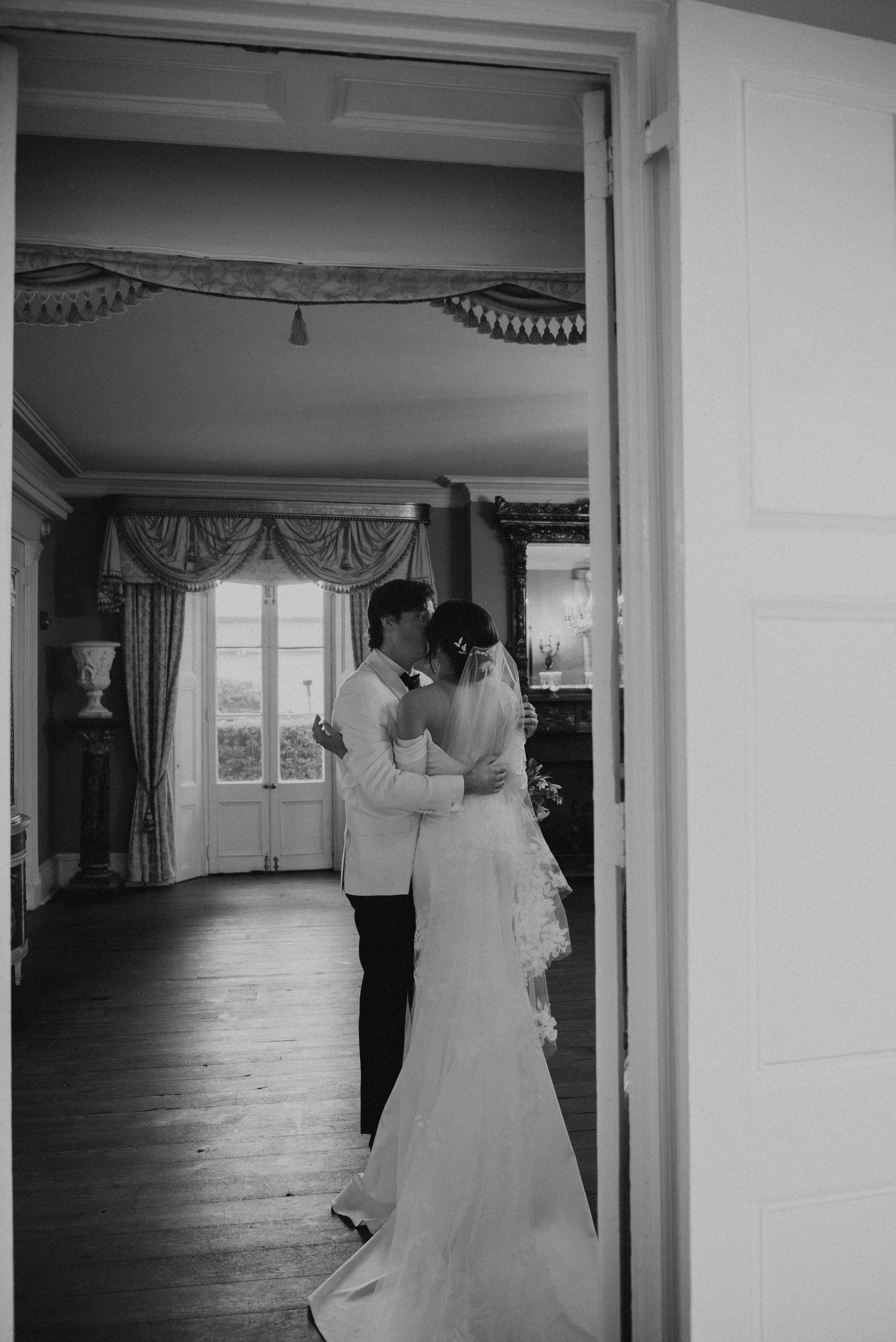 Black and white photo of Bride and groom kissing. Photo is shot through a door way with brides back to the camera.