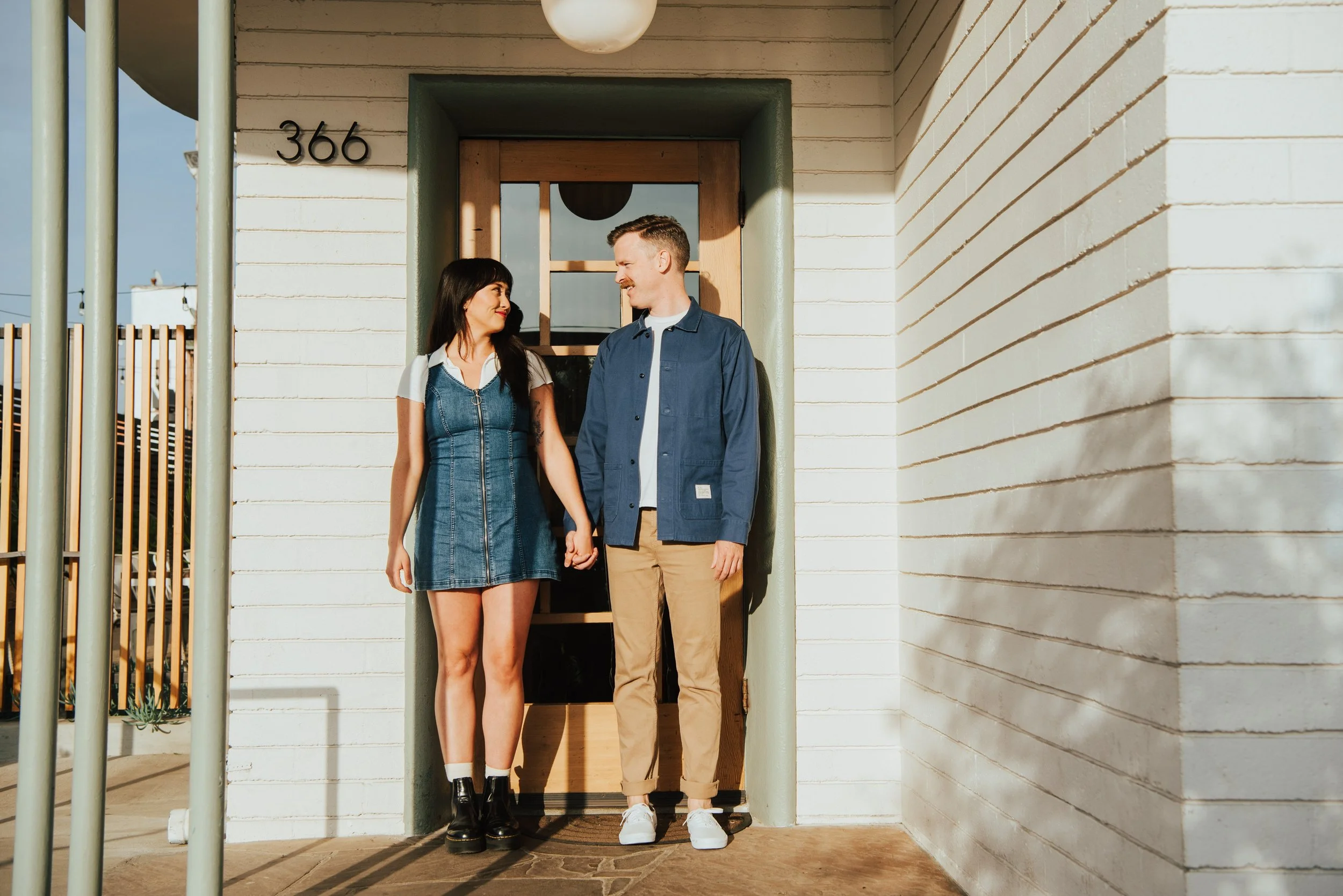 Couple standing in front of a door with their bodies facing the camera but they are looking at each other while holding hands