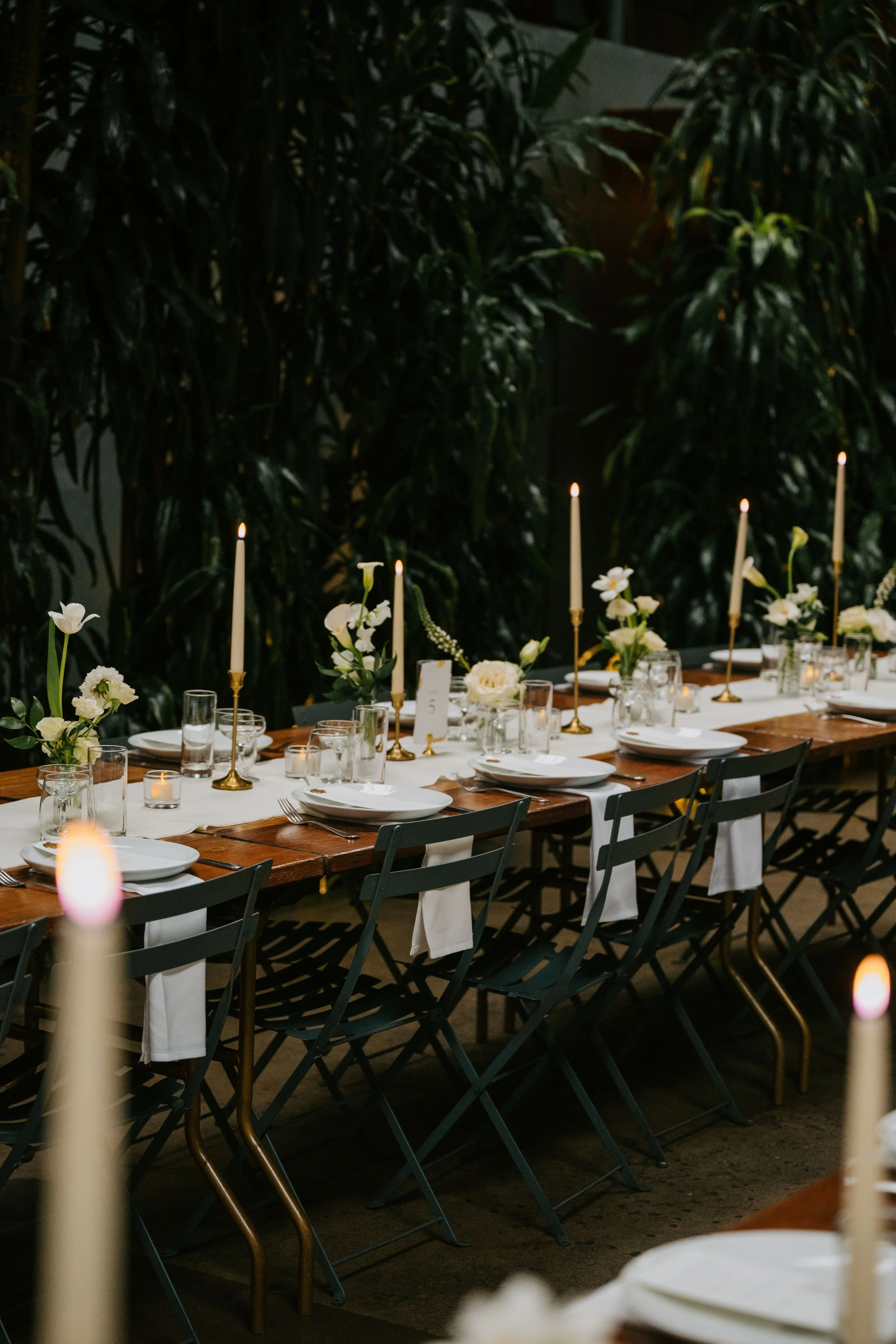 photo of a wedding tablescape with white flowers and candles on the table. A wall of greenery  behind it