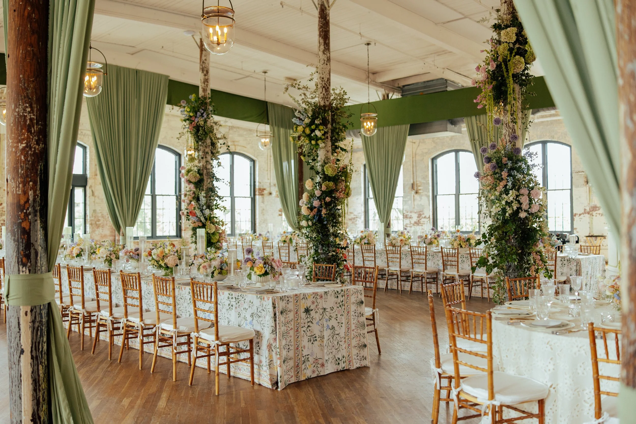 Wedding Reception room all set up with green fabric draped from the ceiling and pastel floral arrangements on tables and around the rooms center polls