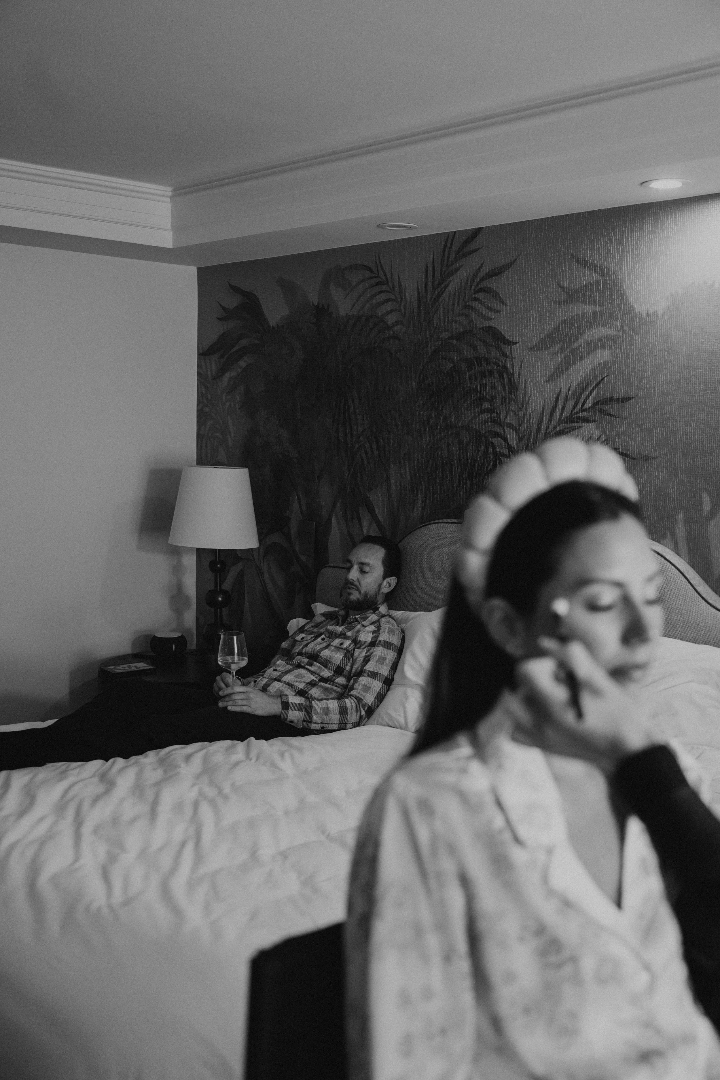 Black and white photo of Groom sitting on bed sipping champagne with bride getting ready in foreground