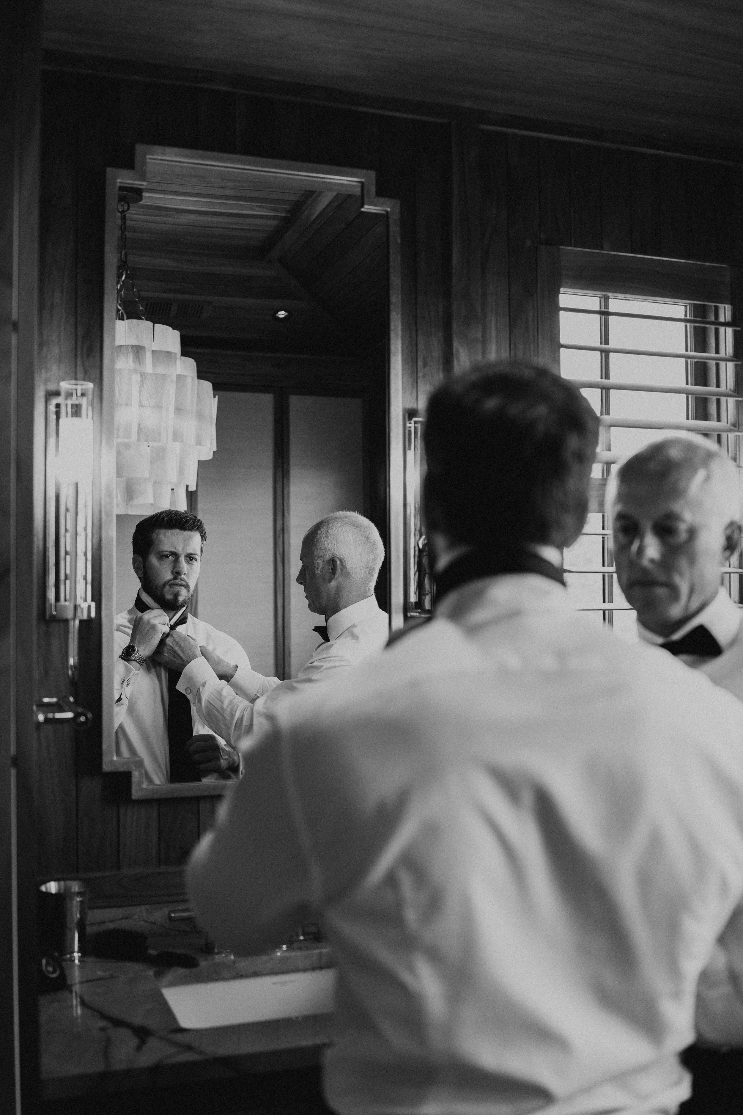 black and white photo of groom looking in a mirror with his dad helping him with his tie