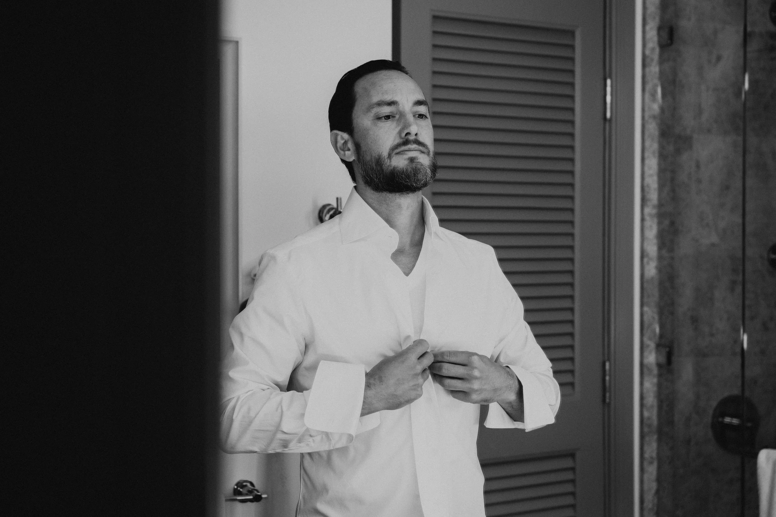 Black and white photo of Groom buttoning up his shirt in the mirror