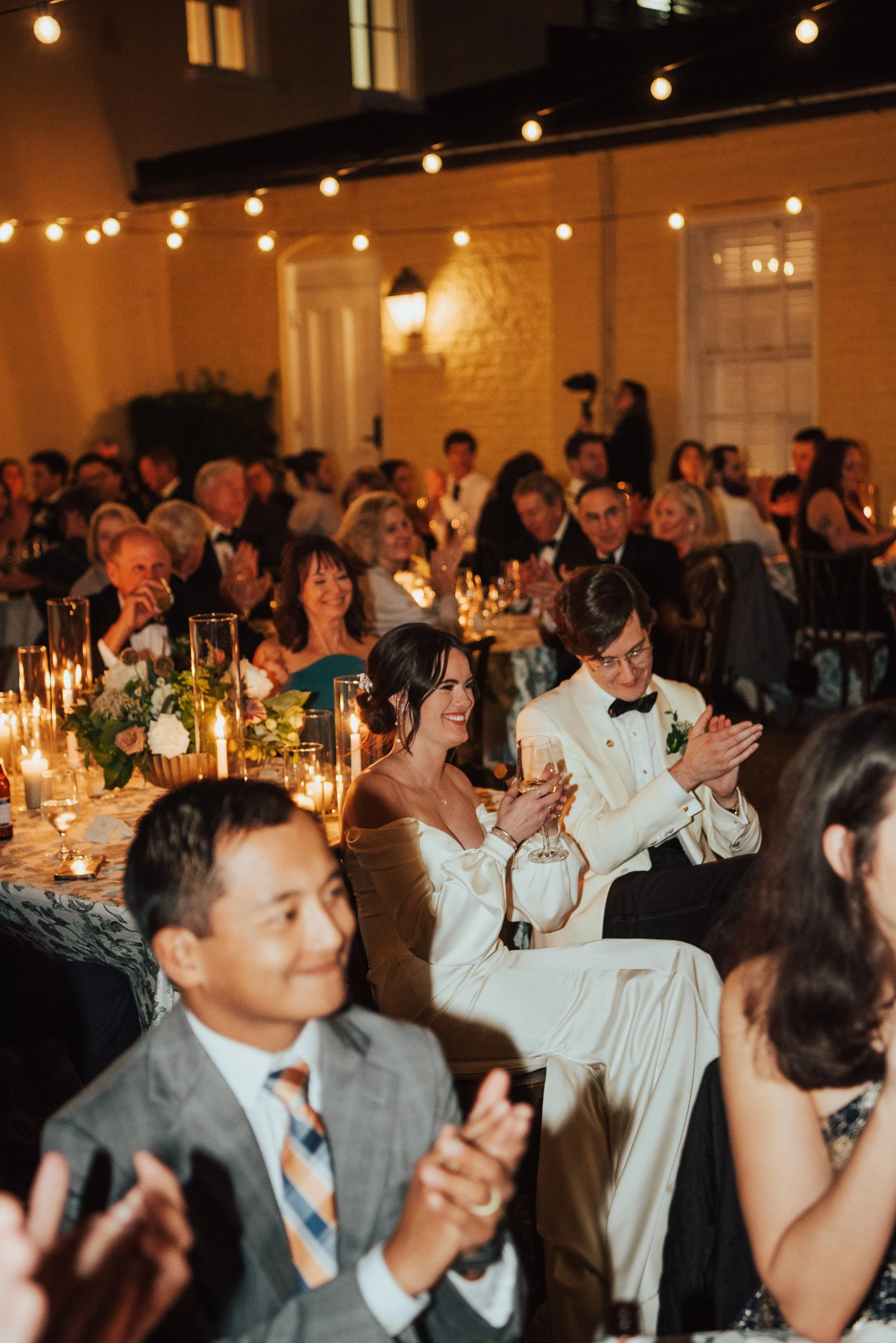 Photo of bride and groom in the midst of their guest at the reception. Everyone is clapping while sitting.