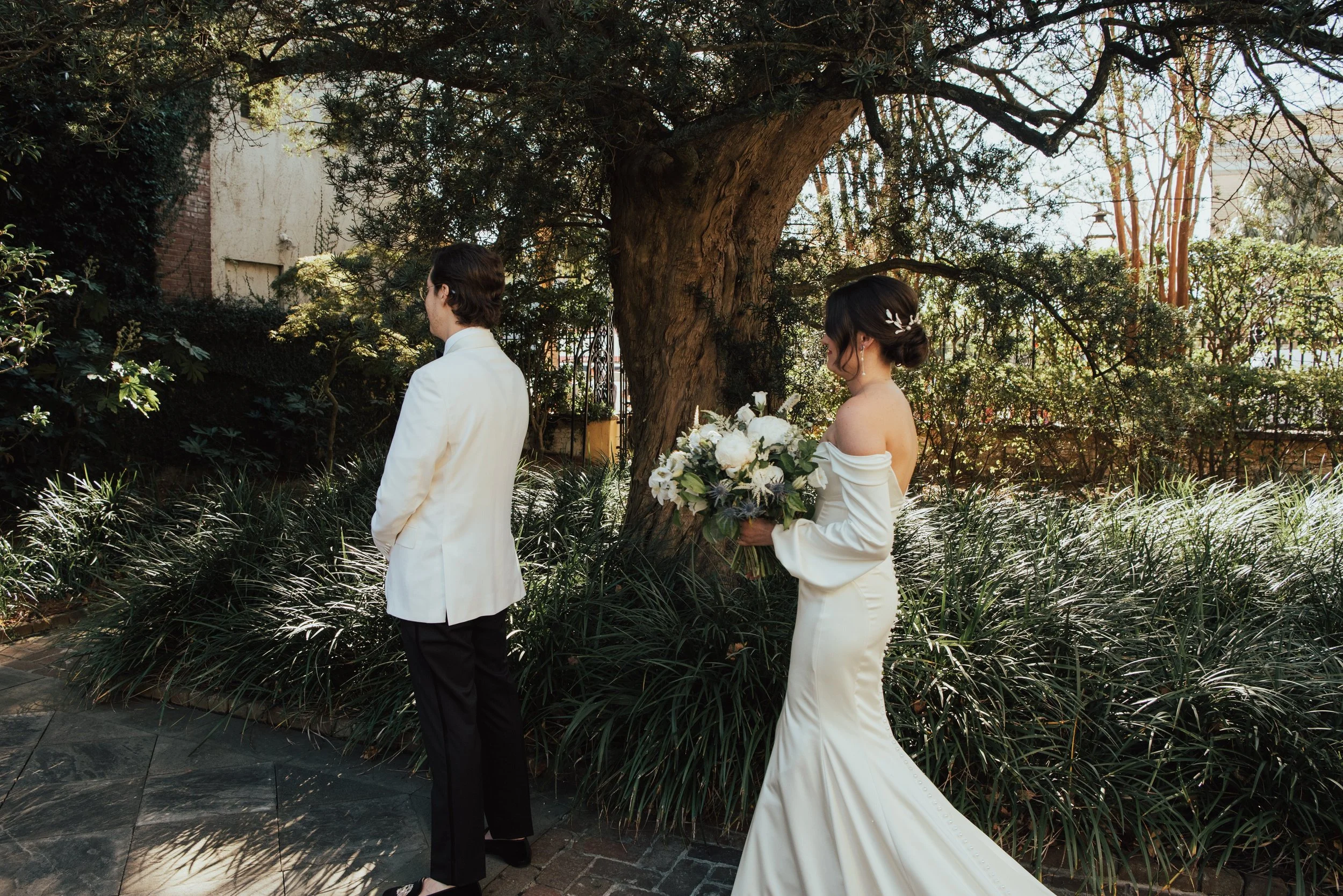 Groom with his back to the camera and his bride is walking up behind him. Shot outside, couple in front of a tree
