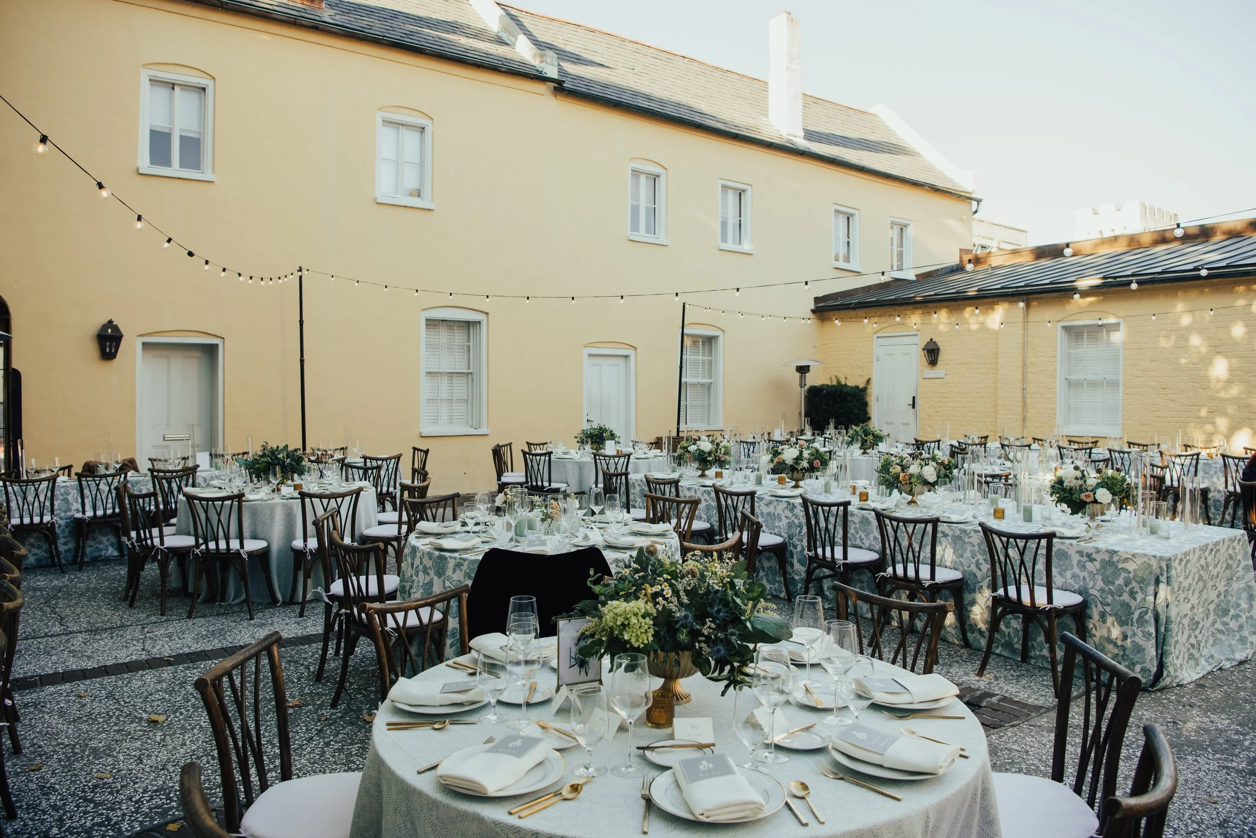 Wide shot of wedding reception set up. The backdrop is a yellow Estate. Bistro lights hang over the tables. The tables are covered by  blue floral patterned table clothes with florals and candles covering the table