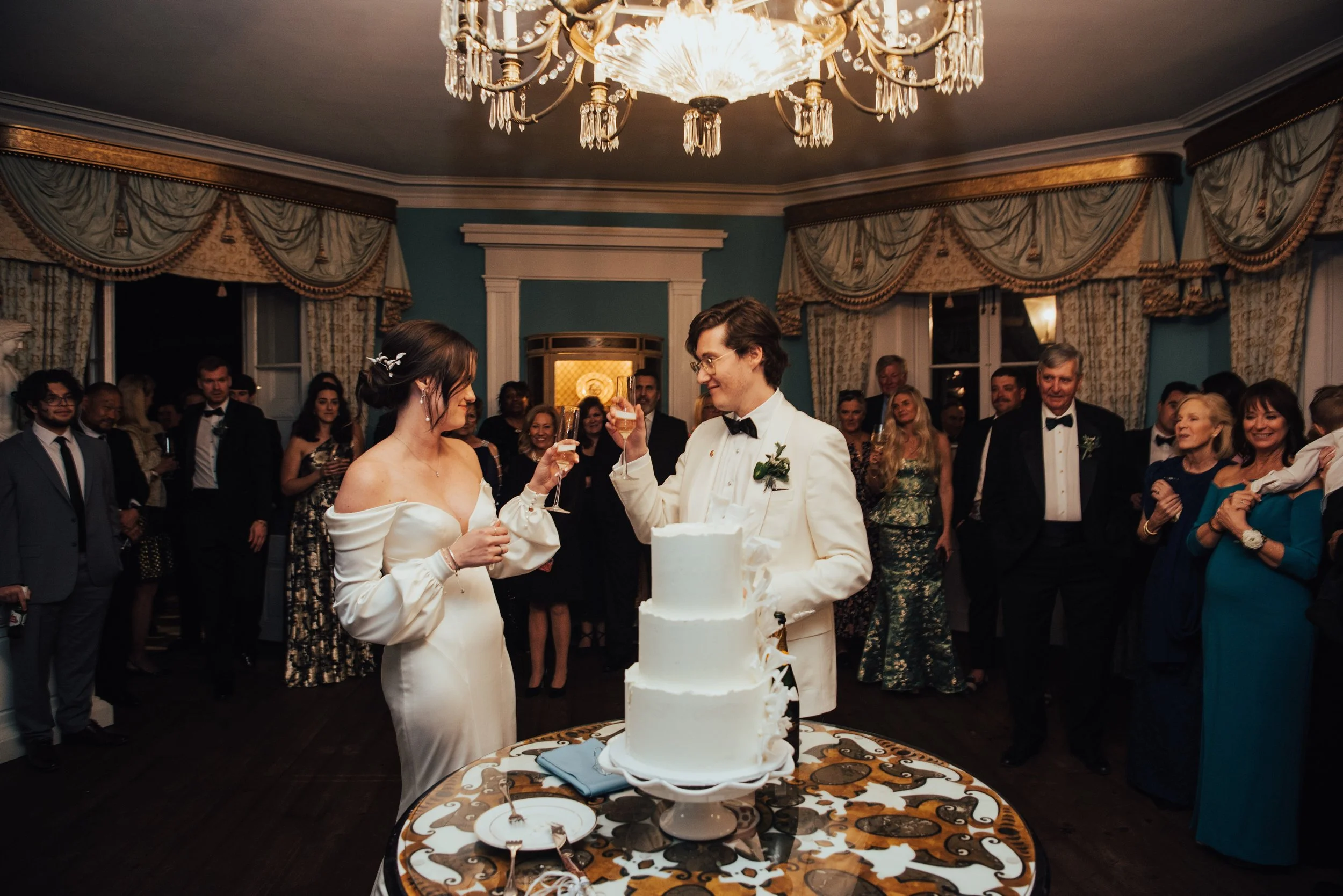 Photo of bride and groom toasting champagne glasses with their guest all behind them. Their wedding cake is in front of them and you can see a chandelier hanging from the ceiling