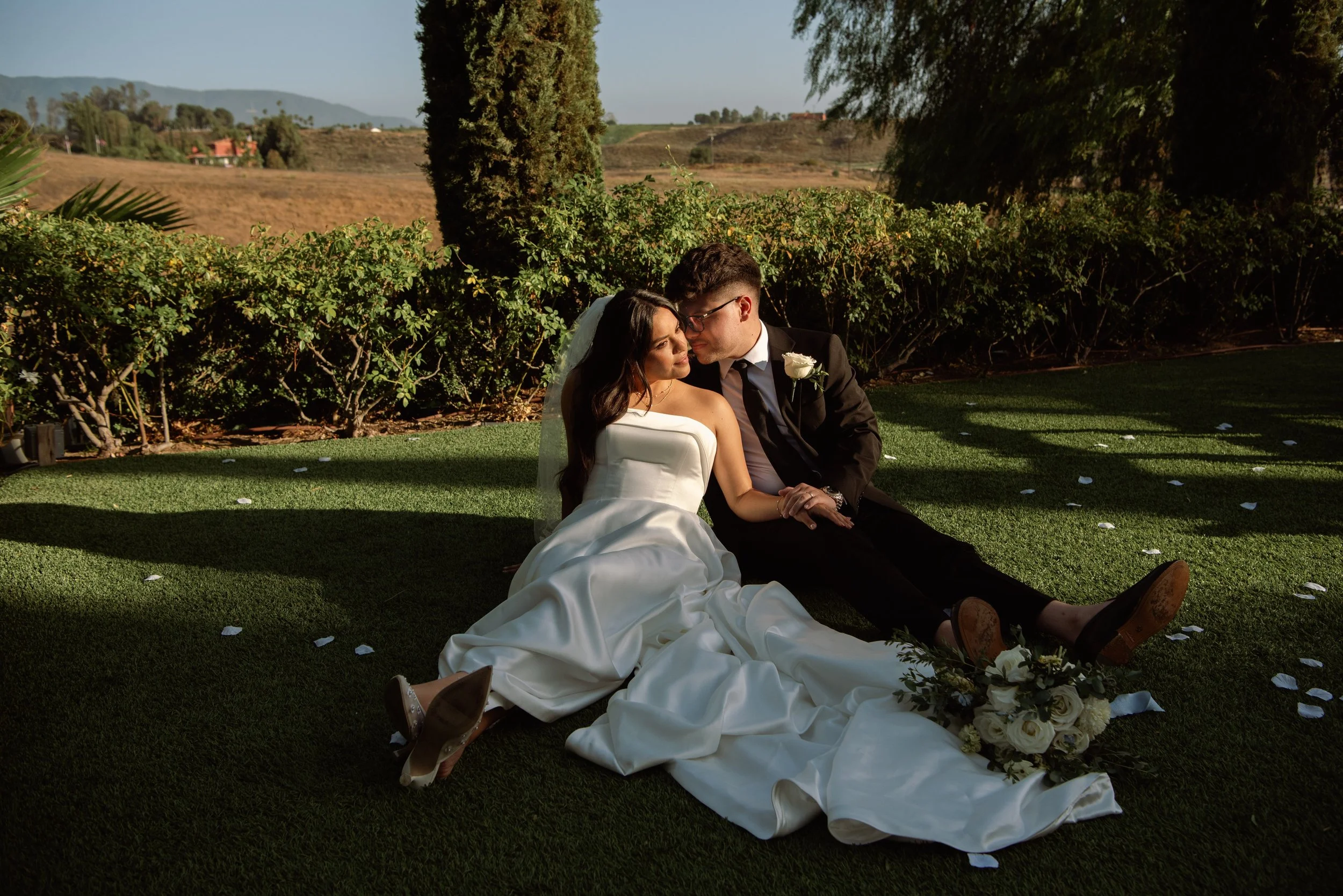 Bride and groom sitting on ground with a patch of sunlight hitting their faces as they lean into each other