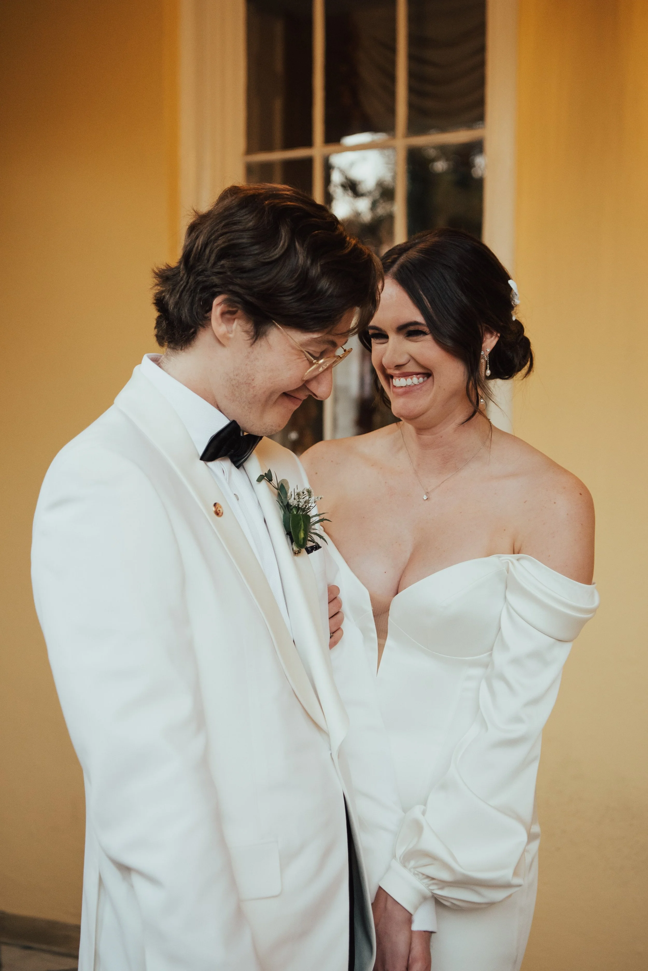 Bride facing camera smiling at groom while hugging his arm. Groom is sideway to the camera wearing a white tuxedo jacket looking down and smiling