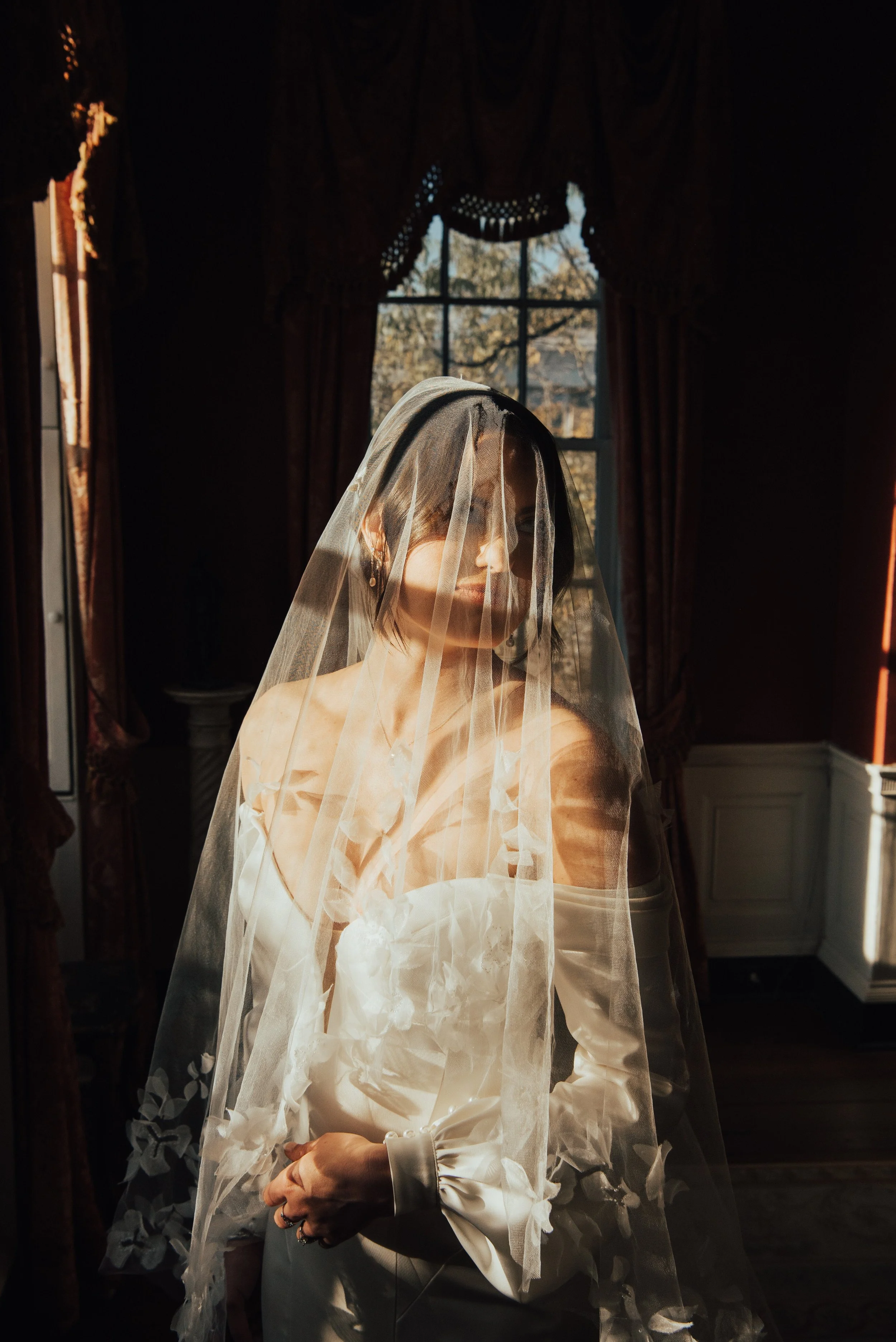 bride standing in front of window. Room is dark as the bride is highlighted by the sun. Bride has her veil over her head