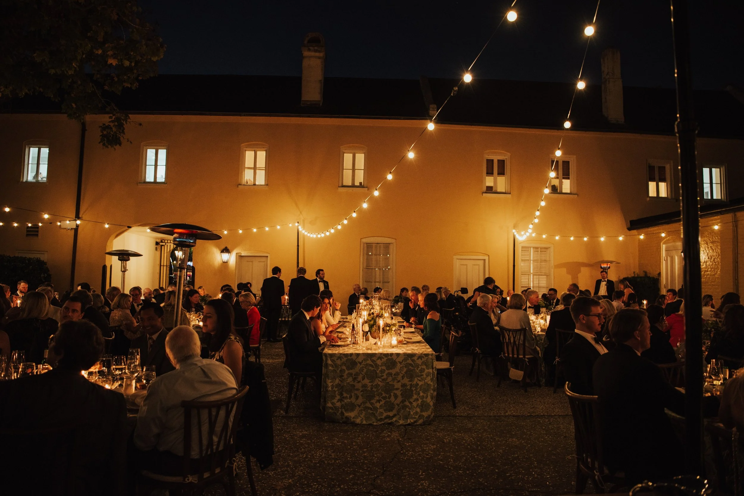Wide photo of Wedding reception Dinner  in a courtyard at night. Gust are sitting at long and round table under bistro lights. The yellow creates an orange glow as the light bounce off of it