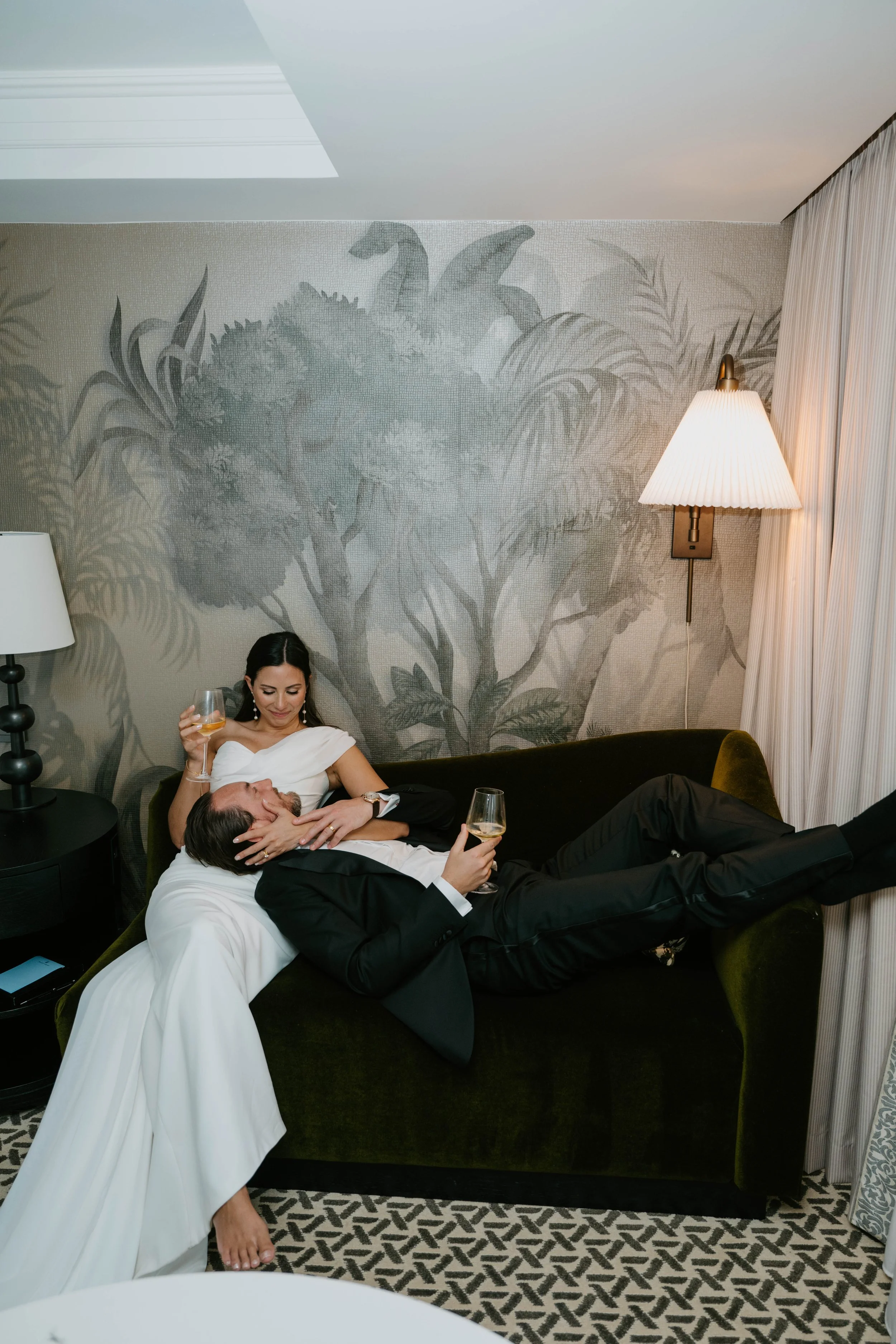 Bride sitting on couch and groom laying on the couch with his head in her lap. Both are sipping champagne