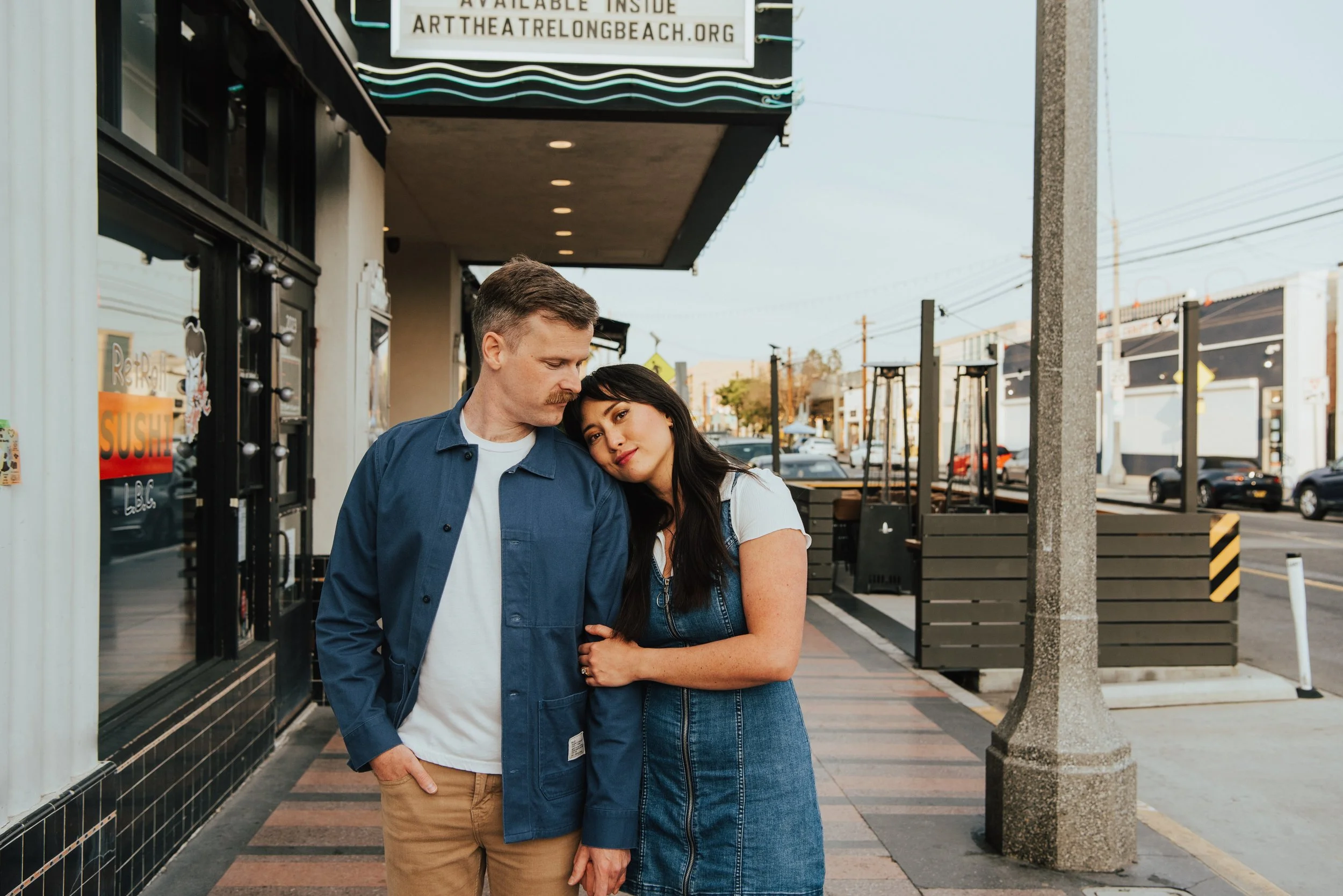 Girl resting her head on guys shoulder and looking directly at the camera and guy looking down towards her.