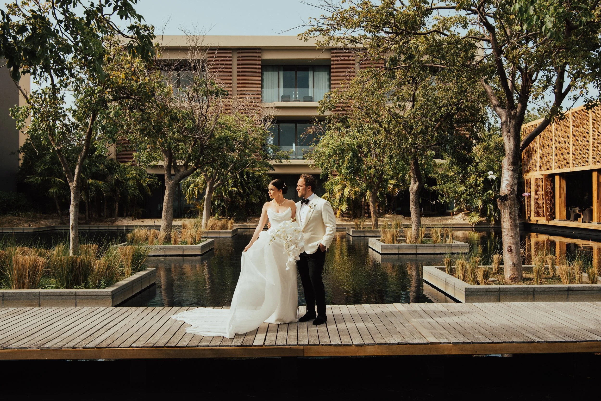 bride and Groom standing next to each other both looking off to the side. The resort with a bunch of trees is behind them
