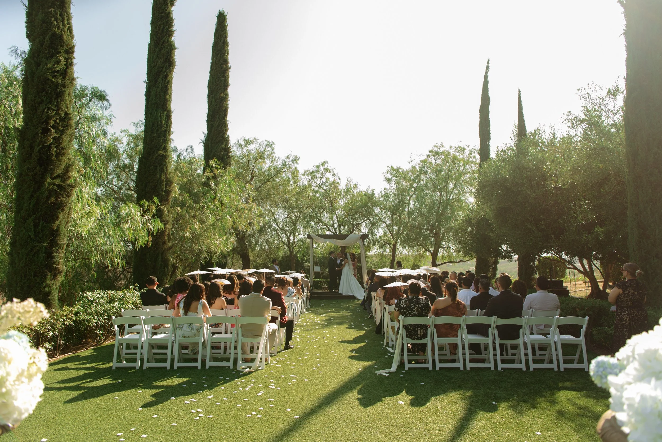 wide shot of wedding ceremony. Ceremony site is surrounded by greenery and Italian cypress tress